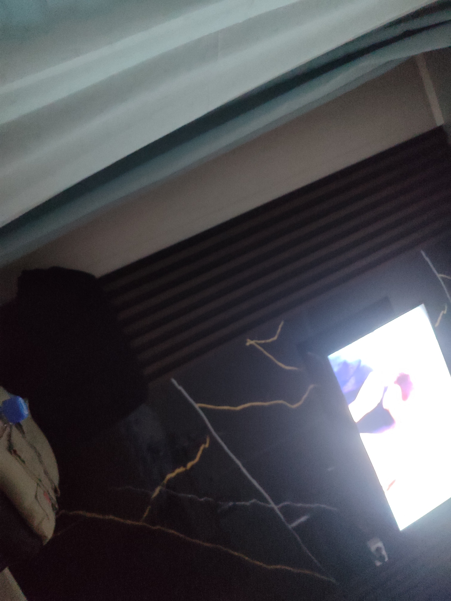 The image depicts an indoor scene, likely a living room. A dark, textured surface, possibly a table with a marble-like pattern, occupies the lower portion of the frame. On this surface, a bright, illuminated screen, likely a television or monitor, displays a pale image with hints of color. To the left of the screen, a dark, amorphous object is visible, possibly a piece of furniture or a person in shadow. In the upper left corner, a translucent, light-colored material, suggestive of curtains, is present. The overall lighting is dim, with the screen being the primary light source. The scene is devoid of any specific landmarks or indicators of its location within Қосшағыл ауылдық округі, Kazakhstan.