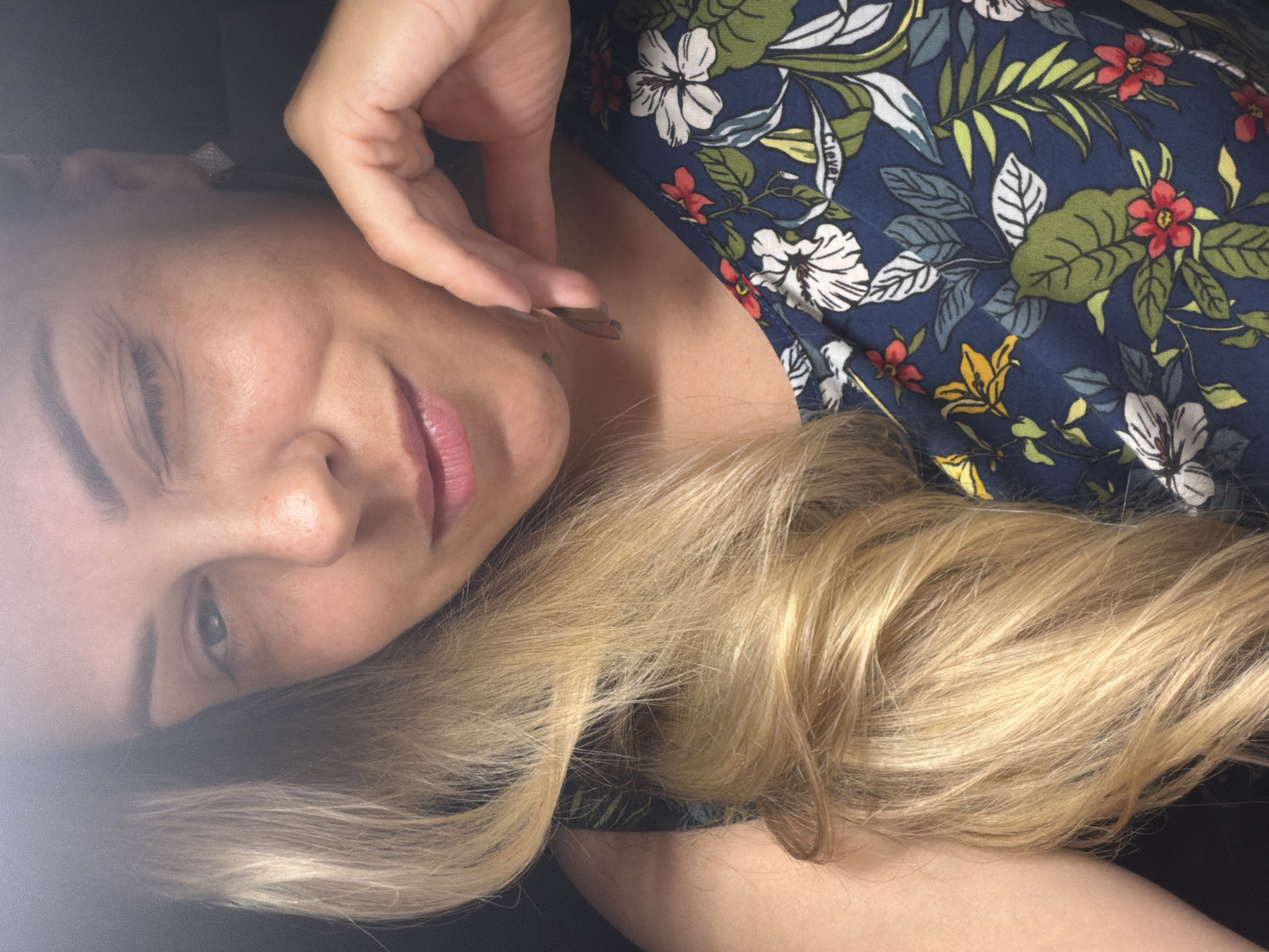 A close-up, high-angle shot features a person lying down, with their head to the left of the frame and their body extending to the right. The individual has fair skin, light blonde hair, and is wearing a dark blue floral-patterned top with white, red, and yellow flowers and green leaves. The person's right hand is near their face, with fingers gently touching their cheek and jawline. Their left eye is visible, looking towards the camera with a soft expression. The lips are slightly parted, revealing a hint of pink lipstick. The lighting is soft, creating a diffused effect, particularly on the left side of the face, suggesting indoor lighting or possibly a slightly overcast day. The background is dark and indistinct, with the focus solely on the person's face, hair, and upper body. The image conveys a sense of relaxation or intimacy. No visible text is present in the image. The location context is Votkinsk, Russia.