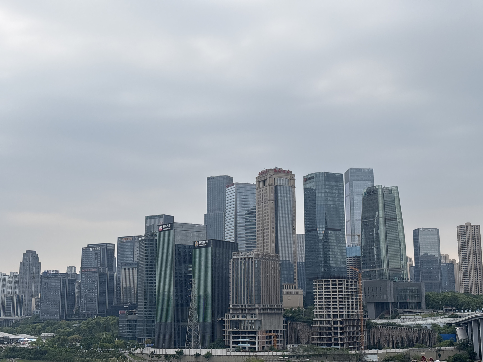 The image depicts a skyline of Yuzhong District, China, characterized by a dense cluster of modern skyscrapers under an overcast sky. The architecture is predominantly high-rise, with glass and steel structures dominating the view. Several buildings are under construction, with incomplete concrete frames visible in the foreground. Vegetation is sparsely distributed at the base of the buildings, near a river. A bridge extends into the scene on the right. No people are visible.