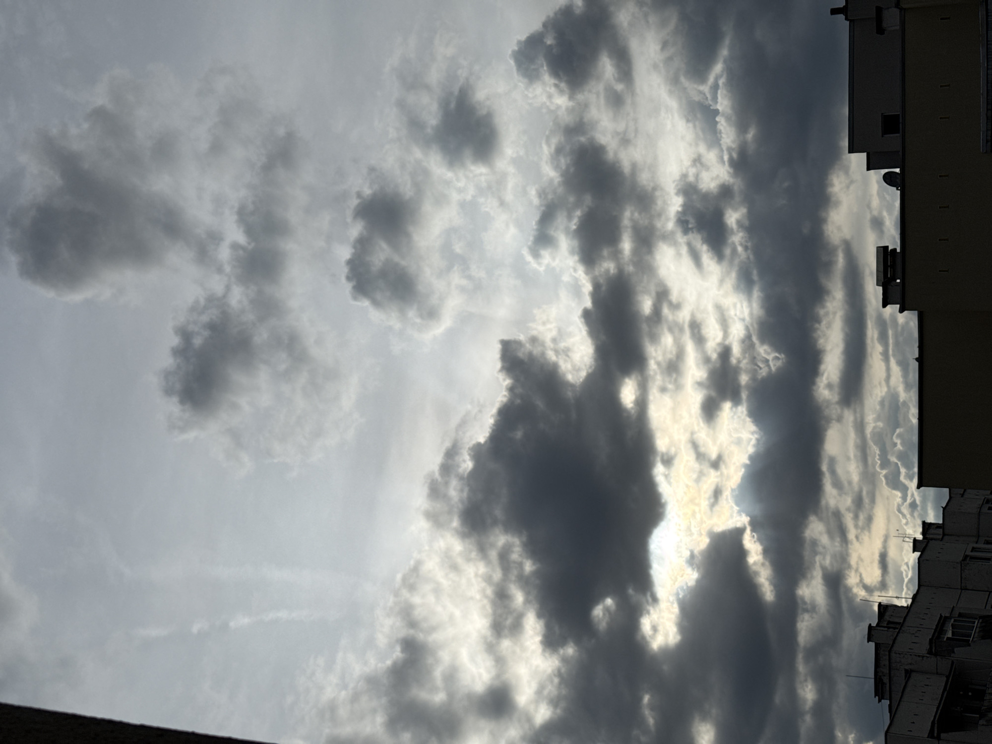 The image shows a cloudy sky over buildings in Pleven, Bulgaria. The sky is filled with dark, heavy clouds, with some lighter areas where the sun is peeking through. The clouds appear to be moving, and the overall atmosphere is dramatic and moody. The buildings below are silhouetted against the sky, and their architecture is typical of urban areas. There are no people or animals visible in the image. The scene suggests an impending storm or a recent downpour.
