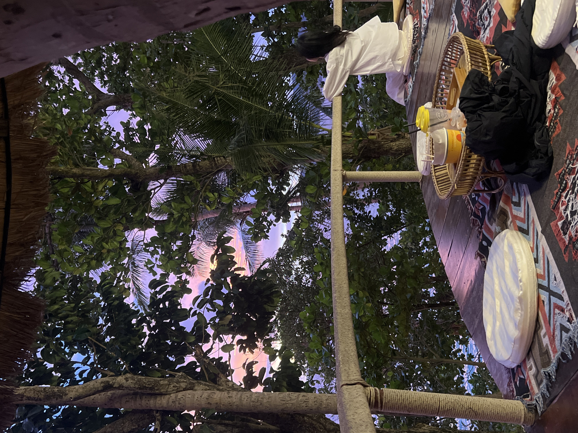 A person in white clothing stands on a wooden deck, looking out at a tropical scene. Lush green trees with palm fronds dominate the view, with a vibrant purple and pink sunset visible through the branches. The sky suggests it is twilight. On the deck, a woven basket holds yellow and white cups with straws, possibly drinks. A black garment is draped over the basket. A patterned rug with geometric designs lies on the wooden planks. A round, white cushion rests on the rug. The scene evokes a sense of relaxation and enjoyment of the natural beauty at dusk, possibly on a balcony or patio overlooking a beach, given the Karon, Thailand location context. There is no visible text in the image.