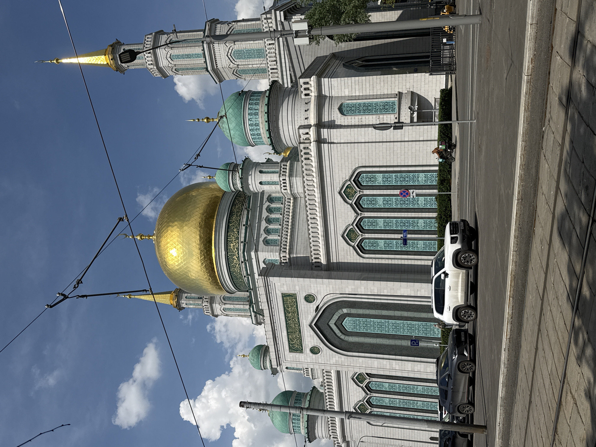 The image shows the Moscow Cathedral Mosque in Moscow, Russia, viewed from street level on a sunny day. The mosque features a large gold dome and several smaller green domes, along with a minaret. The building is decorated with intricate Islamic patterns. Overhead tramlines extend across the sky. In the foreground, a street with parked cars runs alongside the mosque. A person riding a motorbike in traffic is visible in the background. The sky is blue with scattered clouds.