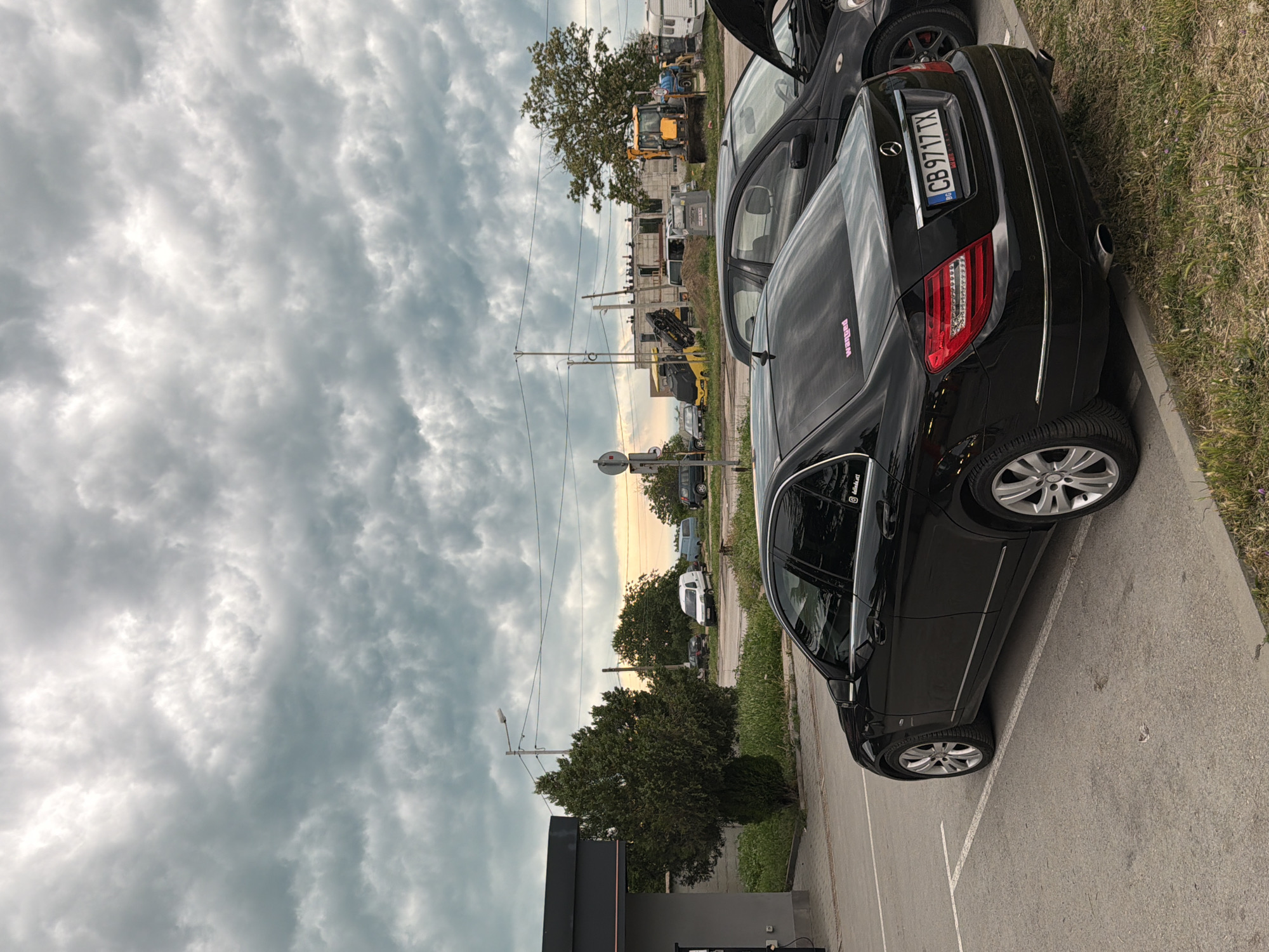 The image depicts an outdoor scene in Varna, Bulgaria, likely during late afternoon or early evening, given the warm, yellowish glow on the horizon under a heavily clouded sky. The weather is overcast, with dense, grey, and textured clouds dominating most of the frame.
In the foreground, a black Mercedes-Benz car is prominently parked on a paved surface marked with white lines. The rear driver's side of the vehicle is visible, showcasing its taillight design, alloy wheels, and chrome trim. The Bulgarian license plate "CB 9717 TX" is clearly readable on the rear. Two stickers are visible on the car: a pink one on the rear windshield, possibly reading "wangan," and a black one on the rear side window, appearing to be "@himati" or similar text. Another dark car is partially visible to the left of the Mercedes.
Beyond the parked cars, a grassy strip and a curb separate the parking area from the background. The midground features what appears to be a construction or industrial site, with a yellow excavator and partially built concrete structures. Various other vehicles, including vans and a camper van, are scattered throughout this area. Utility poles and overhead wires crisscross the scene, with several trees providing greenery. The setting combines elements of urban infrastructure, development, and natural foliage under a dramatic sky.