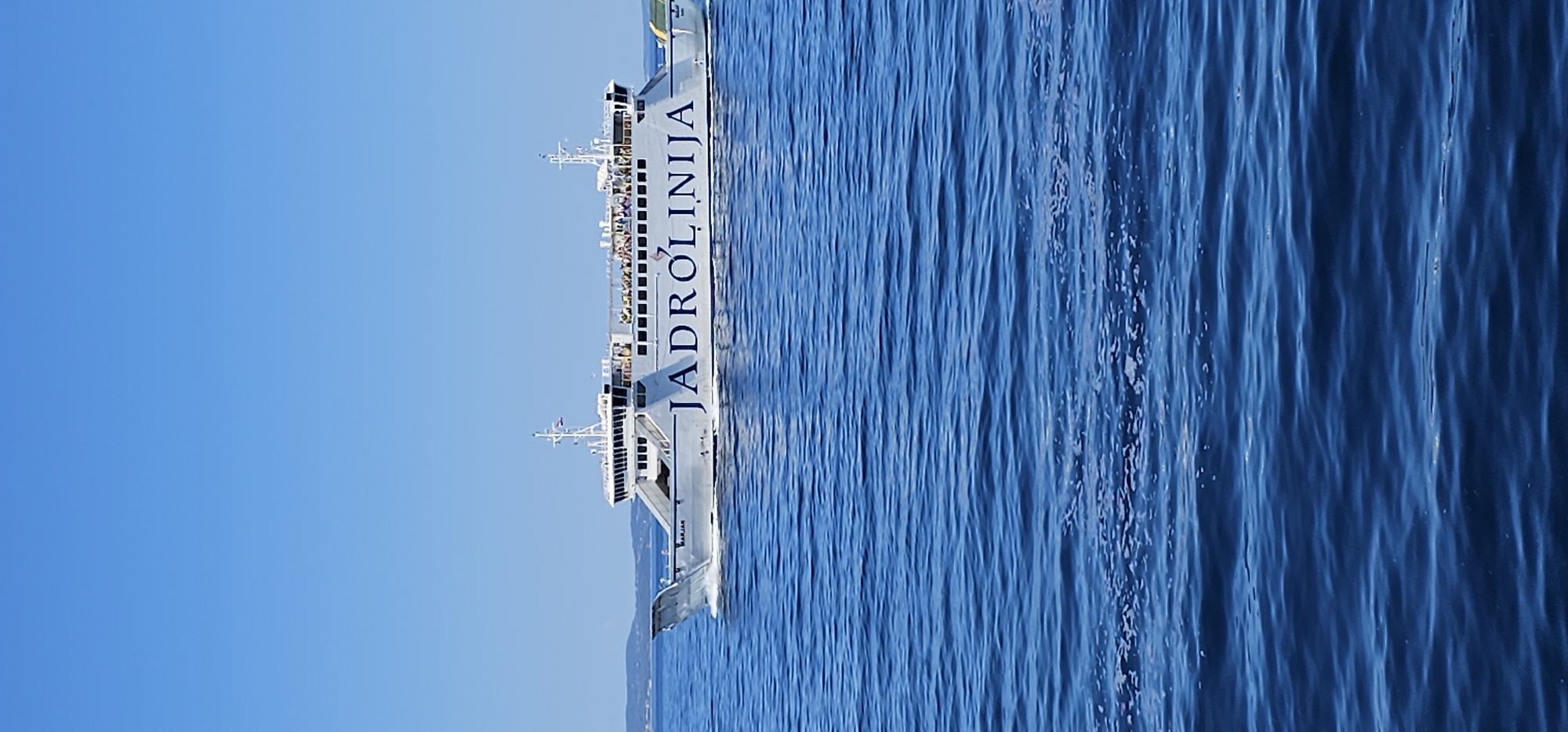 The image shows a ferry with "JADROLINIJA" written on its side, sailing on the sea. The ferry is white with blue lettering and has multiple decks with people visible on them. The sea is calm with gentle ripples. The sky is a clear, bright blue, suggesting a sunny day. In the background, a coastline with hills and vegetation can be seen. The scene appears to be set in a coastal area, possibly a harbor or a route between islands, given the presence of a ferry and the land in the distance. The overall impression is one of travel and leisure on a pleasant day. The time of day is likely daytime due to the bright blue sky and clear visibility.