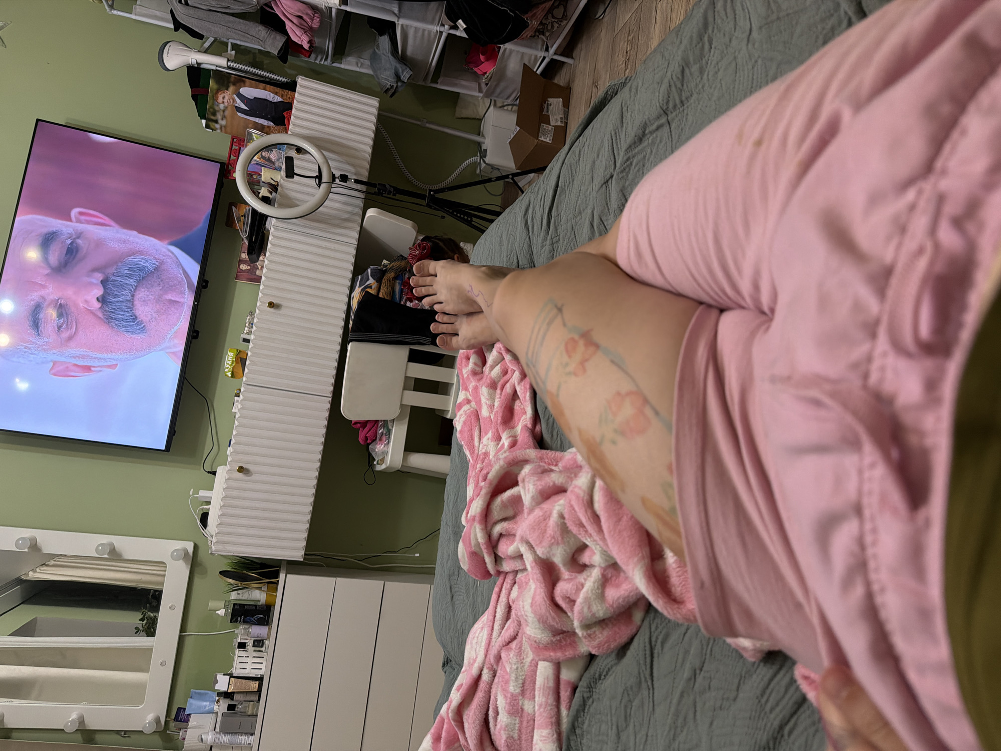 The image shows a bedroom with a person's legs and feet in the foreground, covered by a pink blanket. The person appears to be lying on a bed. In the background, there is a TV screen displaying a man's face, a vanity with makeup and a mirror with lights, a dresser, a chair, and shelves with clothing. The room is decorated in light green and white. There are no visible indications of the location being in Zavetny, Russia.