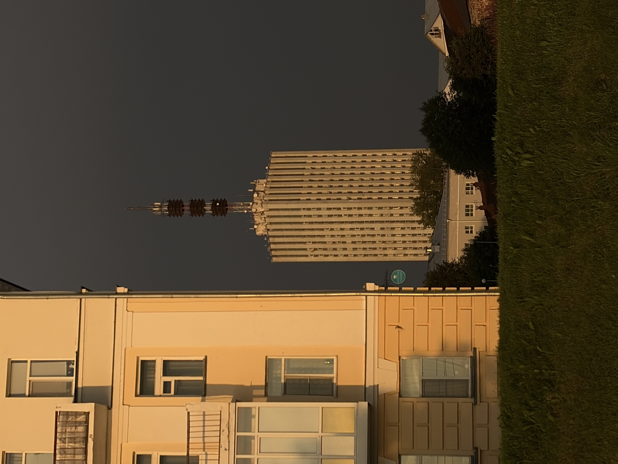 The image shows a television tower in Arkhangelsk, Russia. The tower is tall and cylindrical with a spire at the top. It is surrounded by trees and other buildings. The sky is dark and overcast. The image is taken from a low angle, looking up at the tower. The tower is illuminated by the setting sun, which casts a warm glow on its surface. The scene is peaceful and serene.