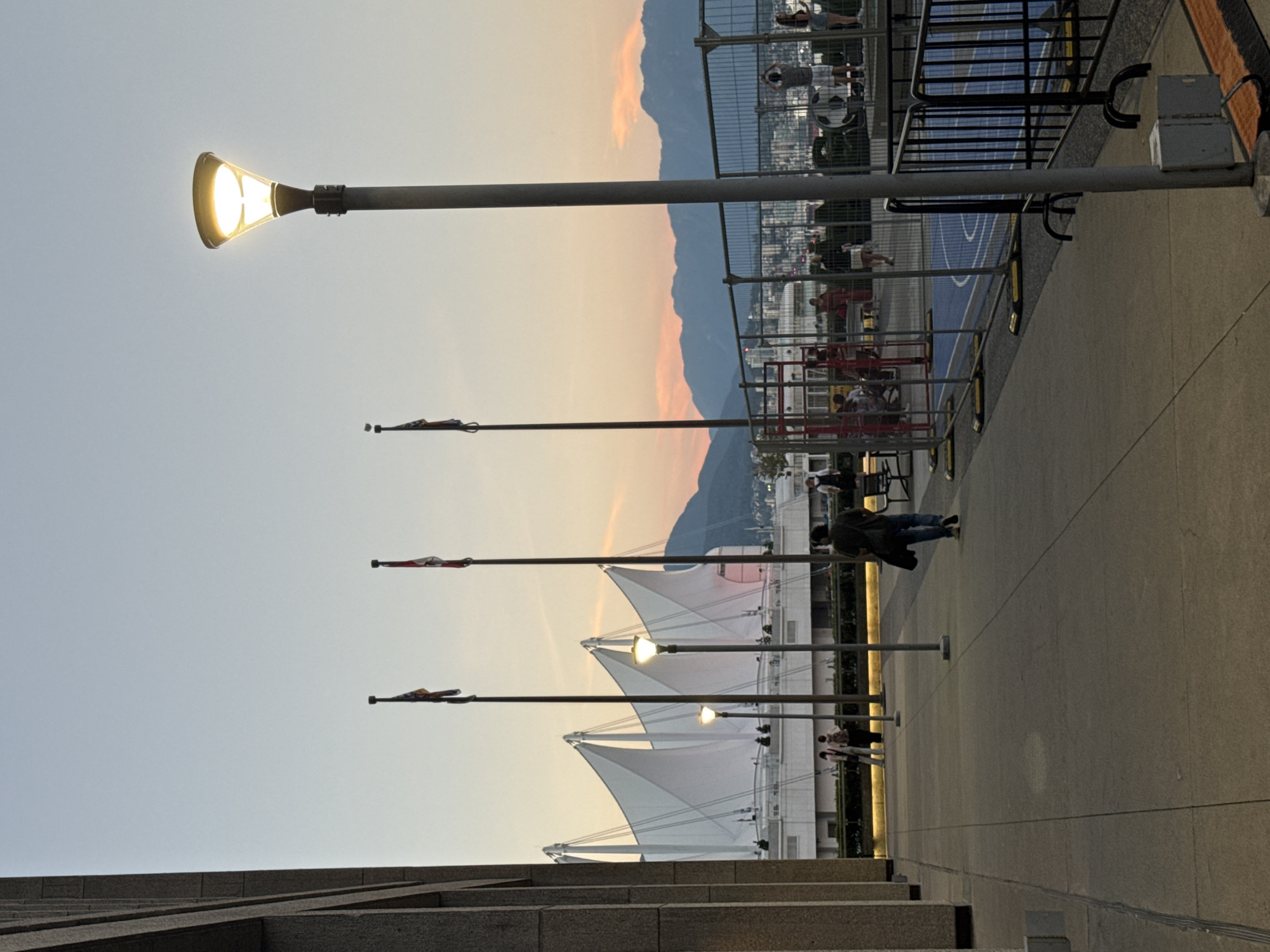 This image depicts a scene at sunset near Canada Place in Vancouver, Canada. The foreground features a concrete pathway with a lamppost casting a warm glow and a section of a building with concrete columns on the left. In the mid-ground, several people are walking and standing, some appearing to be heading towards or away from a fenced-off area that seems to be a court or a play area with blue surfacing. Beyond this, Canada Place's iconic white sails are visible against the dusky sky, with mountains forming the backdrop. The sky exhibits the warm hues of a sunset, with oranges and pinks bleeding into the soft blue. Flags are visible on flagpoles in front of Canada Place. The overall atmosphere suggests an urban outdoor space in the early evening.
