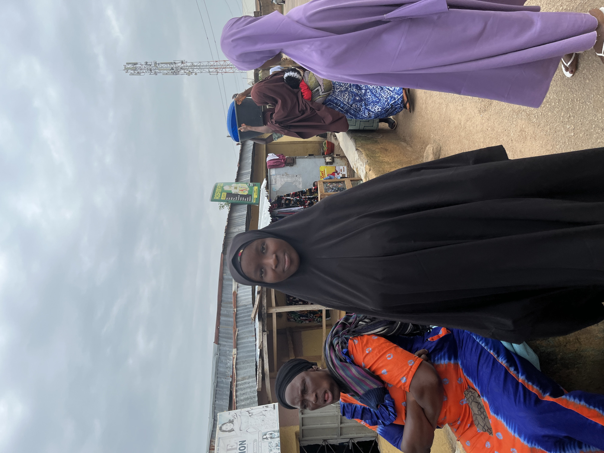 In an outdoor scene in Jos, Nigeria, three women are positioned at varying distances from the camera. The woman in the foreground, facing the camera, is wearing a black hijab and a black garment that covers her from head to toe. To her left, and slightly further back, a woman in an orange and blue tie-dye dress with a patterned scarf around her neck sits with her arms crossed, her face turned to the left and her mouth open. To the right of the foreground woman, another woman is visible from the shoulders down, wearing a light purple hijab and a purple garment, with white sandals on her feet. In the background, other individuals can be seen. One person is carrying a large blue basin on their head, and another is wearing a brown hijab and a blue patterned skirt. The environment includes a building with a corrugated metal roof, a sign with green text, and what appears to be a cell phone tower in the distance. The ground is a mix of dirt and gravel. The sky is overcast.