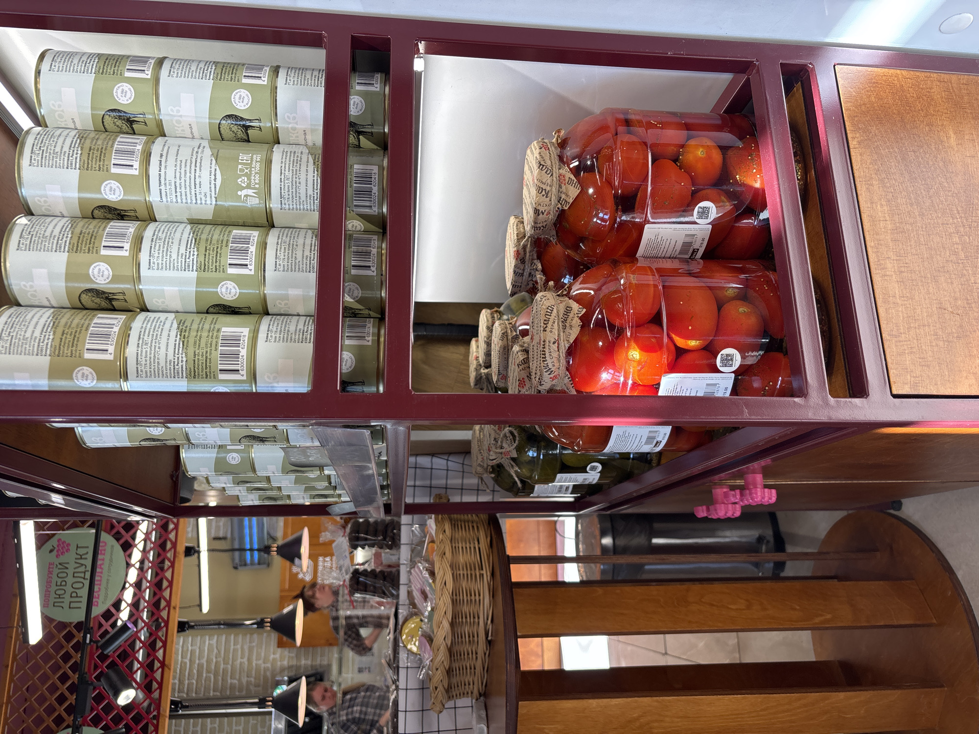 The image shows a retail display of food products in what appears to be a store in Krasnoyarsk, Russia. The shelving is made of dark red metal, featuring several canned goods with labels in Russian. The cans have an image of a bird. Alongside the cans are jars of tomatoes. The tomatoes appear to be a darker red color. The jars are sealed with brown patterned coverings, possibly cloth or paper. Below the canned goods, the image reflects other parts of the store. One sign reads "Try any product... free". In the background, there are lamps, shelves with wrapped products, and the reflections of people. The flooring under the shelving unit is light brown wood. The shelving unit itself has a pink plastic connector that holds the shelves together.