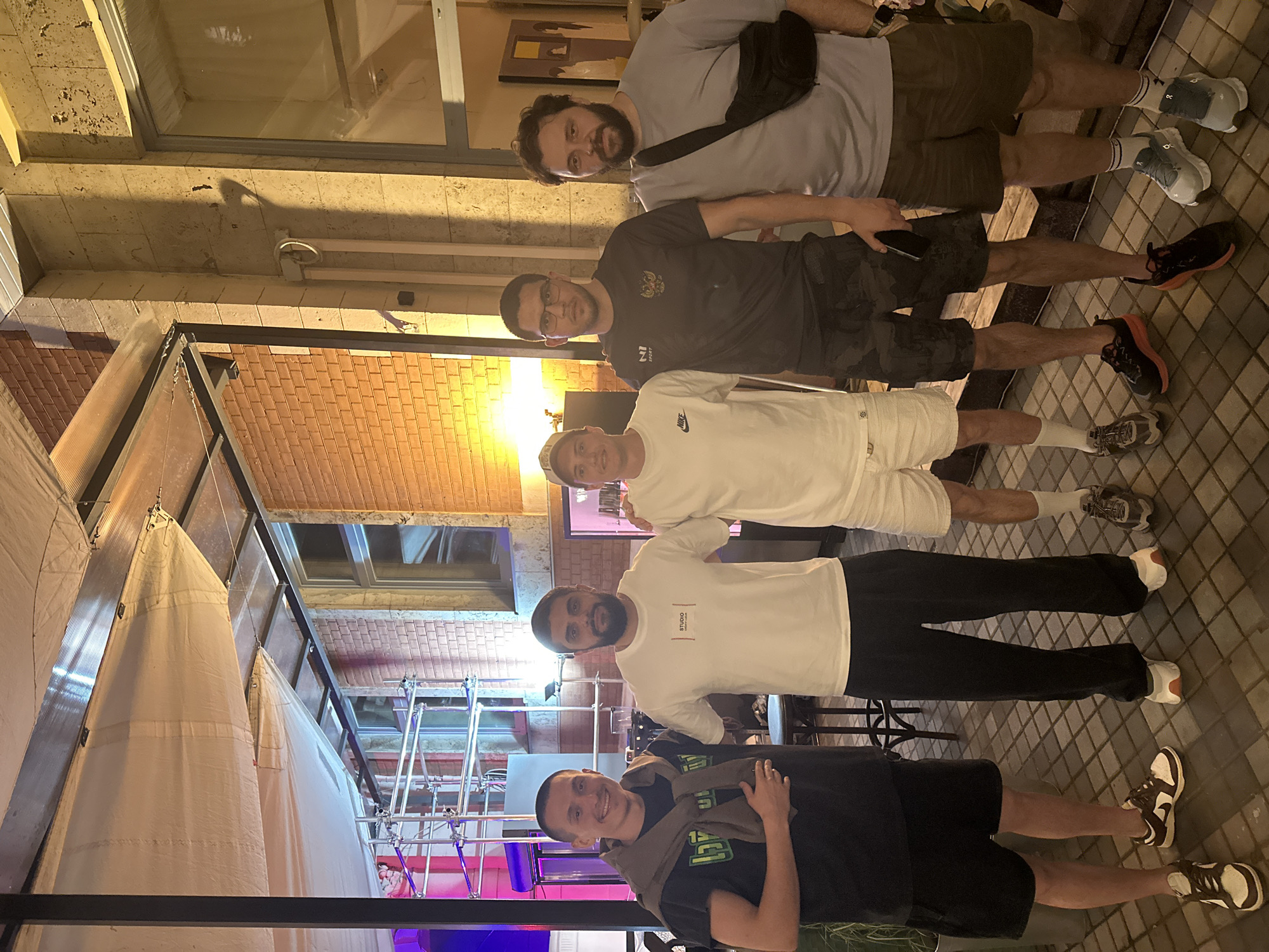 Five men are standing outdoors at night in what appears to be a covered patio area of a restaurant or bar. The men are casually dressed. The man on the far left is wearing a black t-shirt with a brown sweater draped over his shoulders. The second man from the left is wearing a white t-shirt and black pants. The man in the middle is wearing a white t-shirt and white shorts. The fourth man from the left is wearing a dark t-shirt with a crest and camouflage shorts. The man on the far right is wearing a light grey t-shirt and khaki shorts, with a black fanny pack worn across his chest. The background includes brick walls, windows, scaffolding, and a tiled floor. The lighting suggests it is evening. The context of Moscow, Russia, is provided.