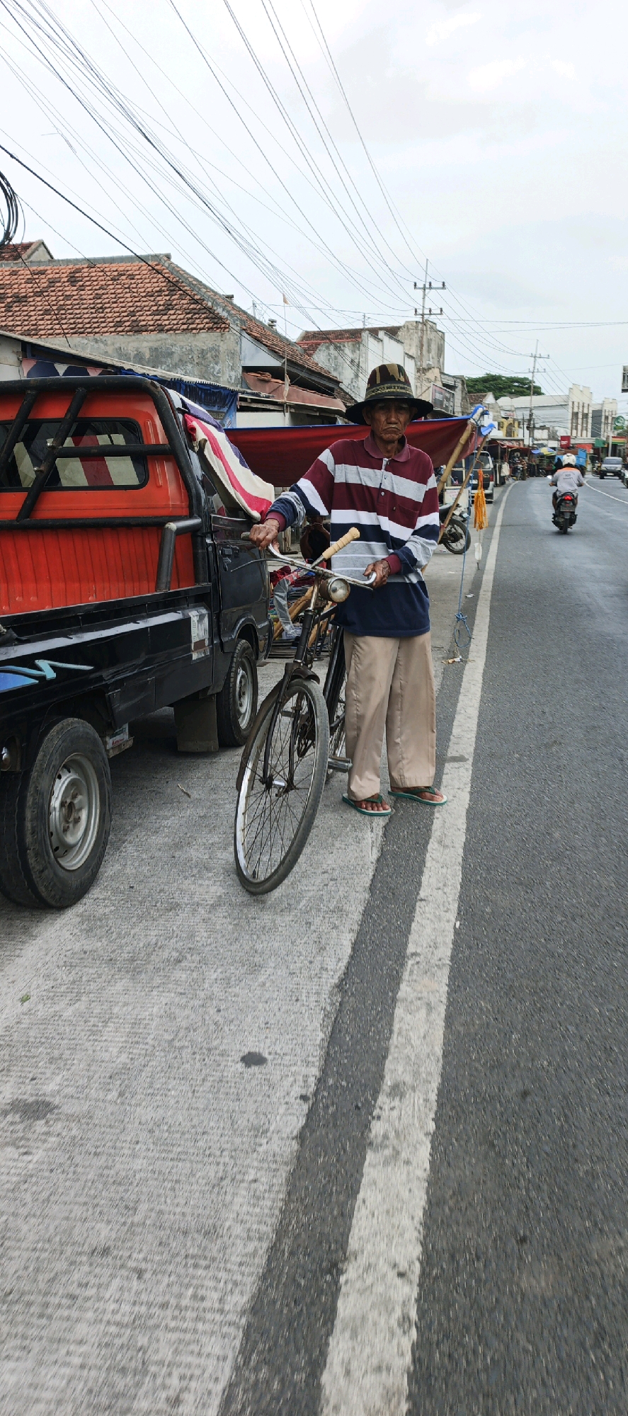 In Kasiyan, Indonesia, an elderly man wearing a striped polo shirt, beige pants, and a patterned hat stands next to a bicycle on a street. The man is holding the handlebars of the bicycle. To his left is the side of a red and black pickup truck. The street has a white line painted on its surface. In the background, buildings with red tile roofs line the street, and traffic, including a motorcycle with a rider, is visible. The sky is overcast.