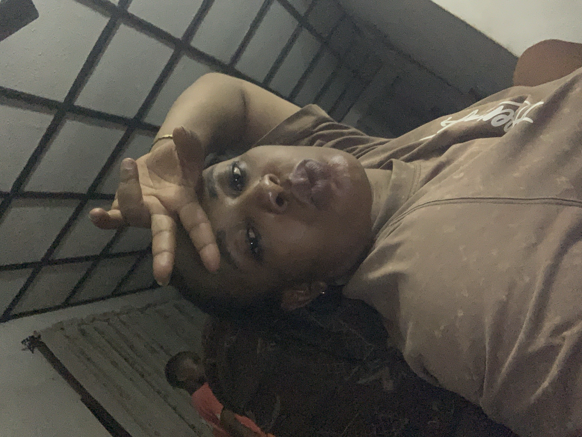 An adult woman with dark skin is prominently featured in a close-up, facing the camera with her lips pursed in a pout. Her right hand is raised, fingers splayed, resting against her forehead and hair. She wears a light brown t-shirt with subtle lighter patterned areas and a white cursive inscription, partially visible, which appears to read "HUSTLERS" on the right side of the chest/sleeve. A small hoop earring is present on her left ear, and a thin gold-colored bracelet is on her left wrist.
In the upper left background, a blurry individual in an orange top is partially visible behind the main subject. The environment is dimly lit, resulting in image graininess. Background elements include a textured, possibly corrugated or planked wall surface on the left. To the right, a dark, grid-like structure, likely a window grill or security gate, is visible against a lighter-toned wall or ceiling. The scene is located in Sabon Tasha, Nigeria, as specified.