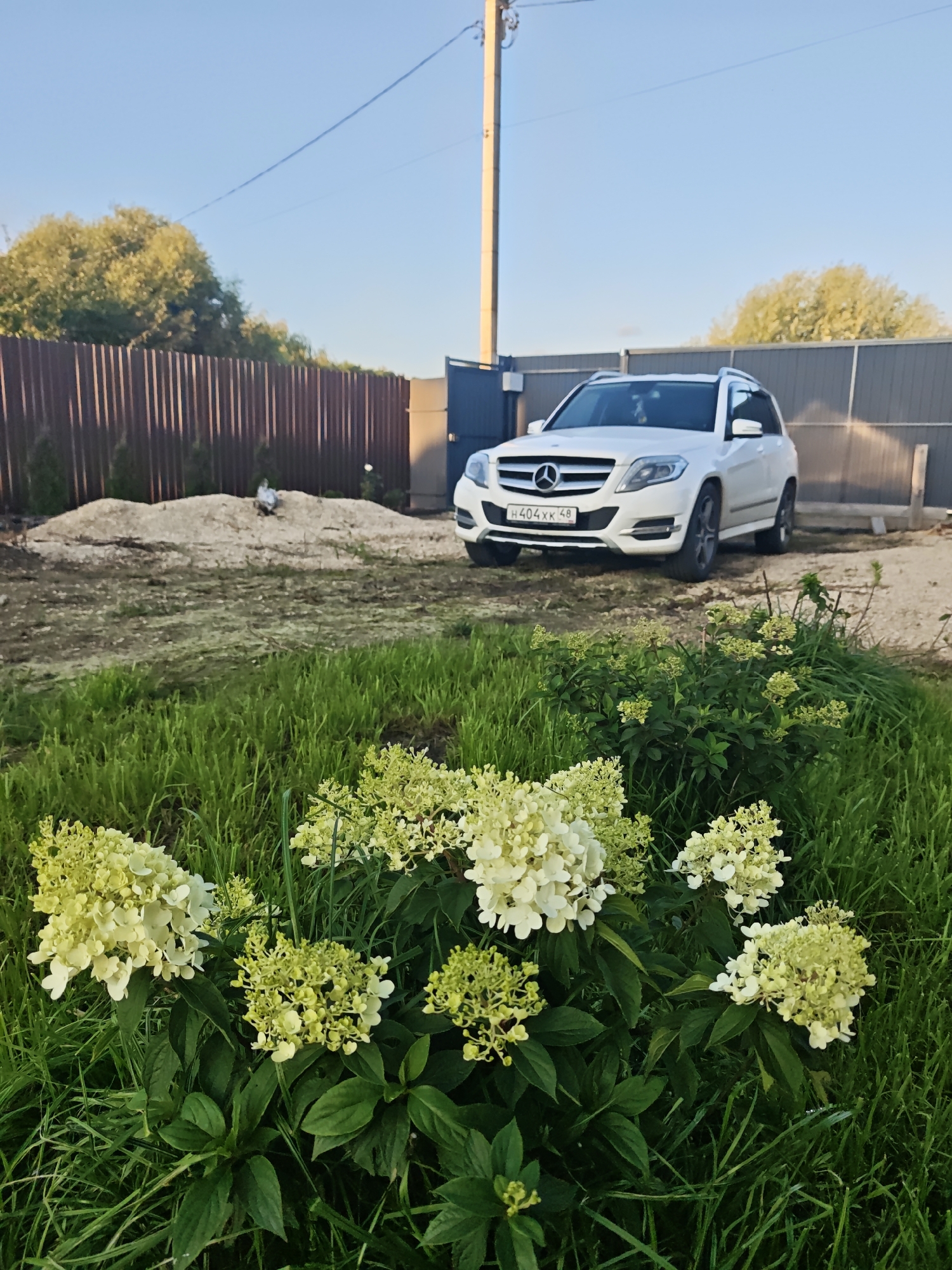 A white Mercedes-Benz SUV is parked on a gravel area in front of a dark brown wooden fence. In the foreground, a lush green grassy area is dotted with flowering plants featuring white and light green blossoms. Behind the fence, a pile of light-colored gravel is visible, and beyond that, trees with green foliage line the horizon. The sky is clear and blue. A tall utility pole stands to the left of the vehicle.