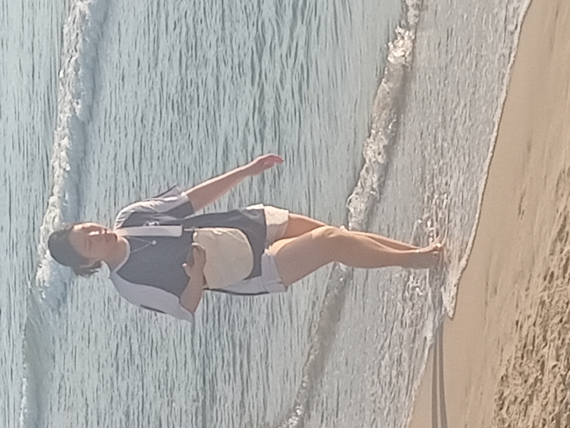 A woman with dark hair tied back is walking along the shoreline at a beach in Da Nang, Vietnam. She is wearing a dark blue and white t-shirt, light-colored shorts, and a cream-colored crossbody bag. Her left hand is holding a mobile phone. The waves are gently lapping at the sandy shore, and the water is a light blue-grey. The lighting suggests it is daytime, possibly late afternoon or early morning. The sand is light brown and appears damp near the water's edge.