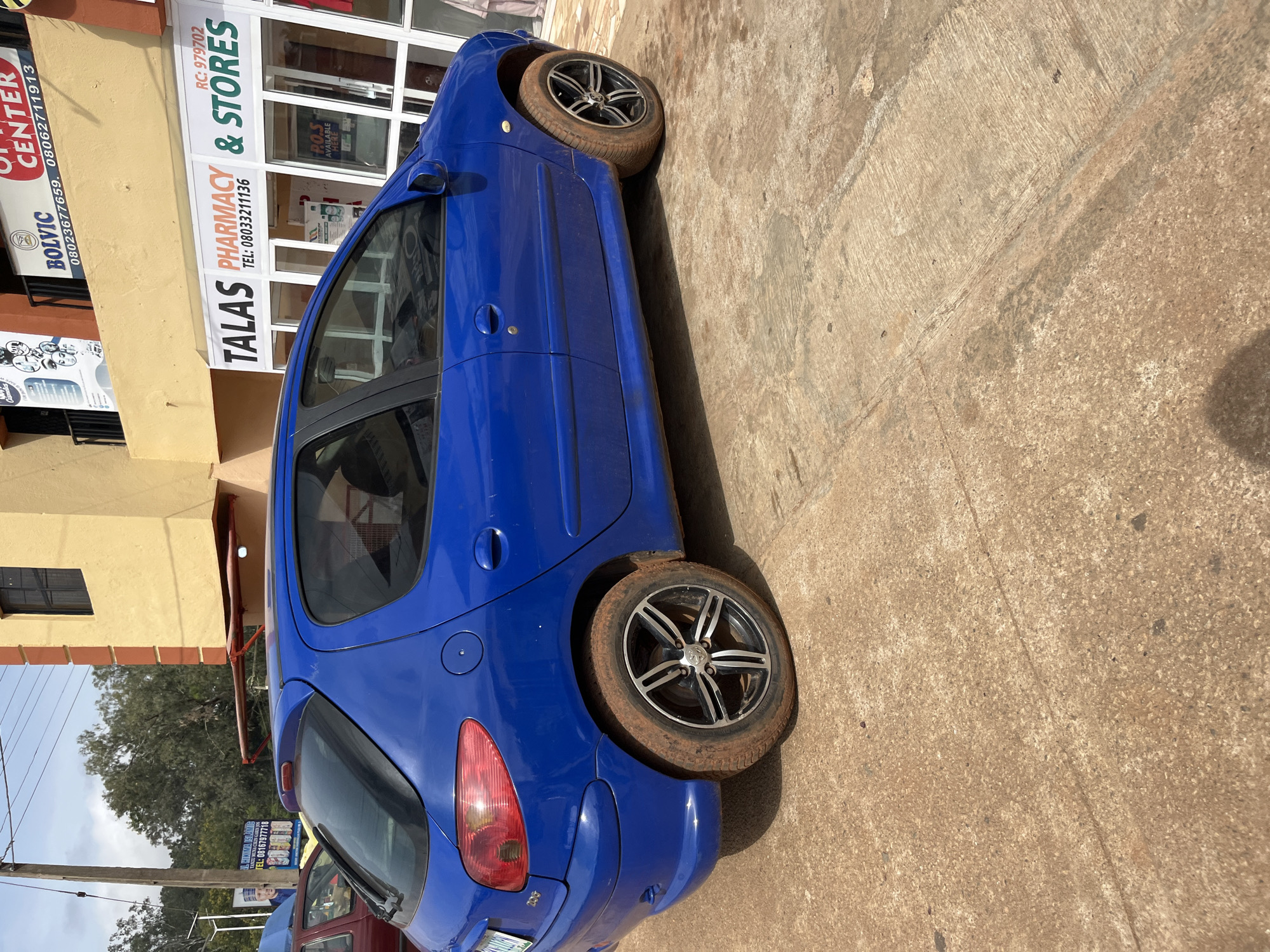 In Bukuru, Nigeria, a blue Peugeot 206 hatchback is parked on a dirt-covered concrete surface in front of a building. The car's tires are caked with reddish-brown mud. The building has a yellow facade with orange trim and a large sign that reads "TALAS PHARMACY & STORES". Above this sign, another sign advertises "BOLVIC". The sky is overcast.