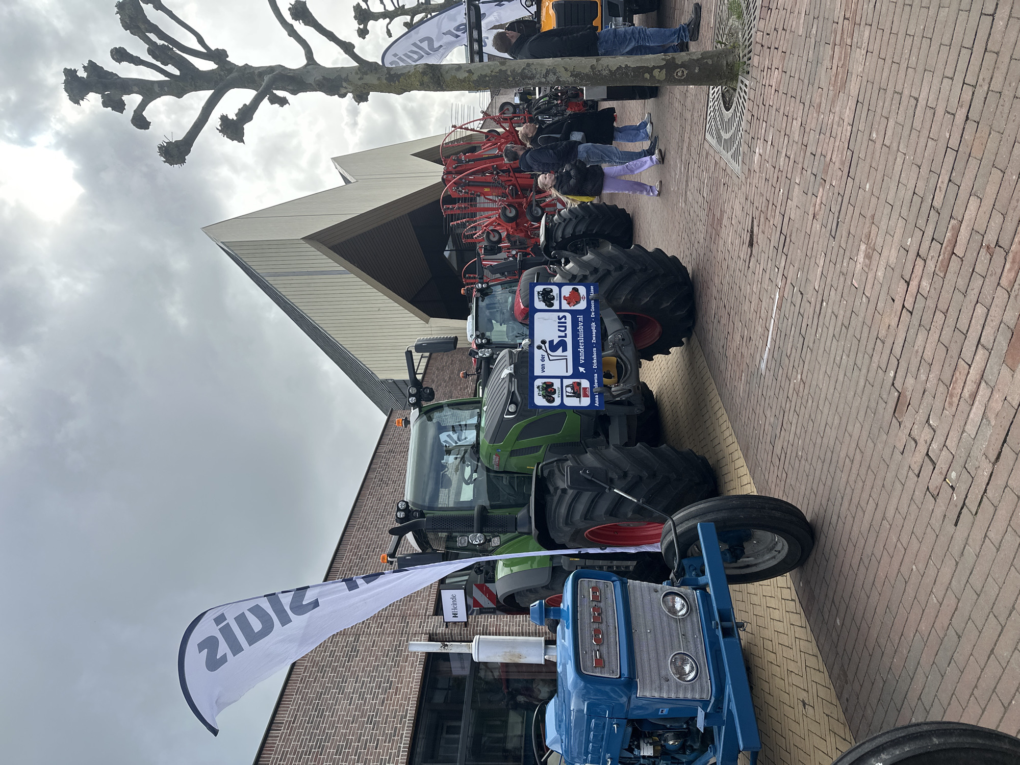 The scene depicts an outdoor agricultural machinery display on a brick-paved public area in Schagen, Netherlands. The specific location is in front of the Markt 18 building, distinguishable by its modern angular architecture with light-colored paneling, and an adjacent multi-story brick building featuring a "Heinde" sign in a ground-floor window. The sky above is uniformly overcast.
Several agricultural vehicles are exhibited. In the foreground on the left, an older blue Ford tractor with a silver grille and dual headlights is present. Adjacent to it, a large, modern green Fendt tractor with prominent black tires and red wheel rims is on display. A blue sign on the front of the green tractor identifies "van der Sluis" and lists "vandersluisbv.nl" along with several regional Dutch locations (Anna Paulowna, Dirkschorn, Zwaagdijk, De Goorn, Lisse). Behind these, large red agricultural implements, consistent with hay rakes or similar machinery, are visible. Portions of additional tractors, including a red model and a yellow/orange model, are discernible further into the display area on the right.
Approximately six to eight individuals are present, observing the exhibited machinery. One young person, wearing light purple pants and a dark jacket, is walking through the mid-ground. Several adults in casual attire are standing in small groups, positioned to view the equipment. A mature, pollarded tree with a thick trunk and gnarled, bare branches stands on the right side of the paved area, partially obstructing the view. The ground is paved predominantly with reddish-brown bricks laid in a herringbone pattern, with some lighter-toned paving sections near the blue tractor. White teardrop-shaped banners, bearing dark, partially reversed text, are positioned near the exhibits.