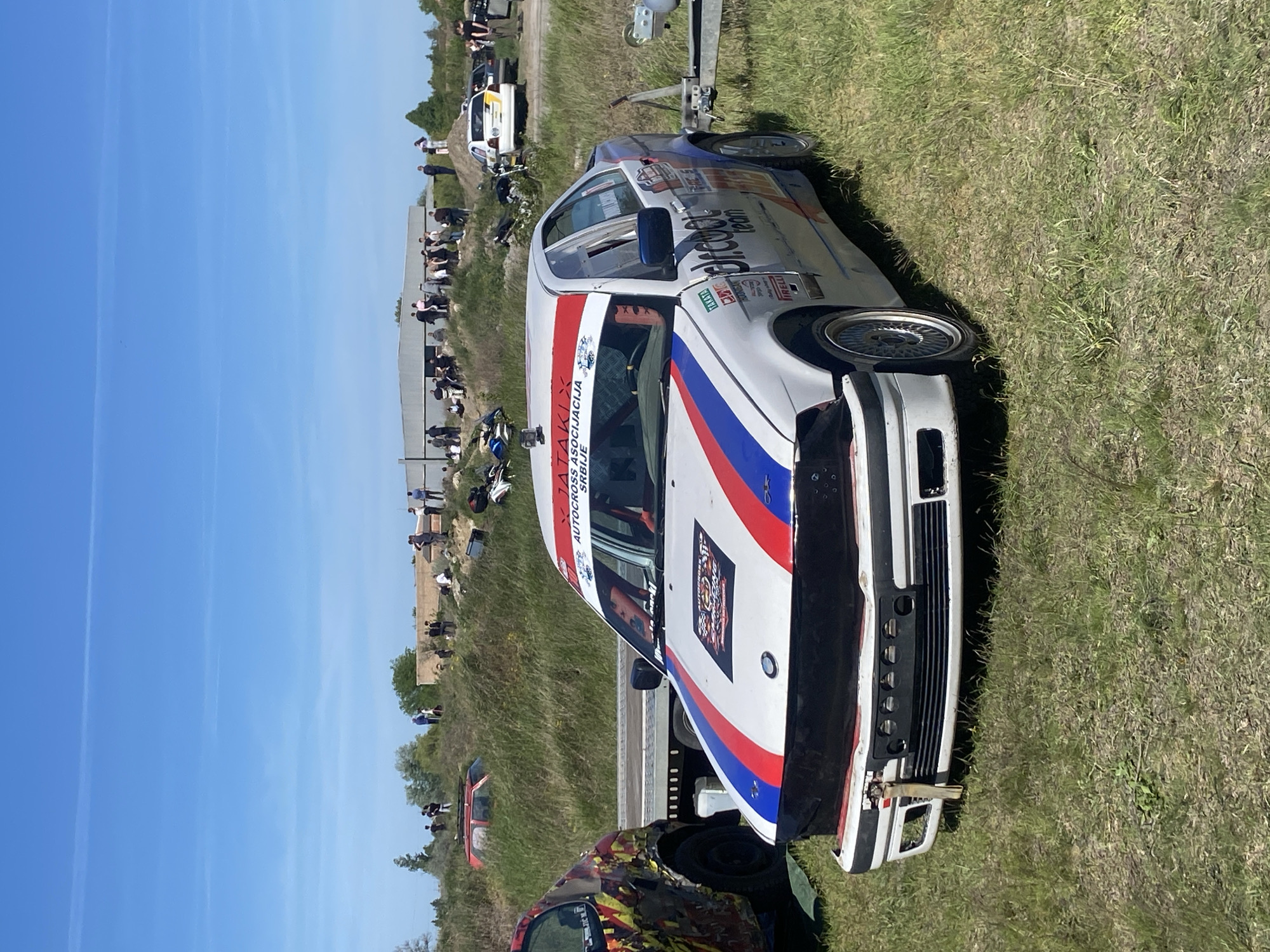 The image depicts an autocross event in the Kanjiza Municipality, Serbia. A white race car with red, white, and blue striping is in the foreground. The car has sponsors' logos on its sides and hood. A driver is visible inside. A crowd of spectators stands behind a barrier along the edge of the track. Another race car is on a trailer in the background. The sky is clear and blue. The event is taking place in a grassy field.