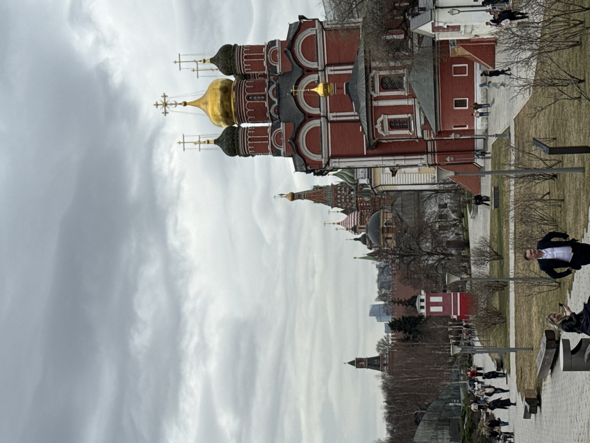 An outdoor scene in Zaryadye Park, Moscow, Russia, depicts the historic Church of the Conception of Saint Anna under an overcast, light grey sky. The church, positioned on the right, is constructed of red brick with white decorative trim, featuring a large central golden onion dome flanked by four dark-domed towers, all topped with golden Orthodox crosses. A smaller golden dome with a cross is visible on a lower section. In the background, the red brick walls and towers of the Moscow Kremlin are discernible on the left, with the distinctive multi-colored domes of St. Basil's Cathedral further to the right of the Kremlin. Modern skyscrapers are faintly visible beyond the Kremlin walls.
The foreground and midground consist of a paved walkway winding through a park area with dormant deciduous bushes and trees. Modern streetlights with dual lamps are regularly spaced. A small, red-and-white striped rectangular building stands in the mid-left park area.
Numerous individuals are present. In the right foreground, a man in a dark suit jacket and white shirt stands with his left hand on his hip, looking towards the right. To his left, a blonde woman is seated on a low concrete bench, looking down at a smartphone. Groups of people are walking along the main paved path in the mid-left background and near the church structure on the right. All individuals are dressed in casual to smart-casual attire, suitable for cooler weather.