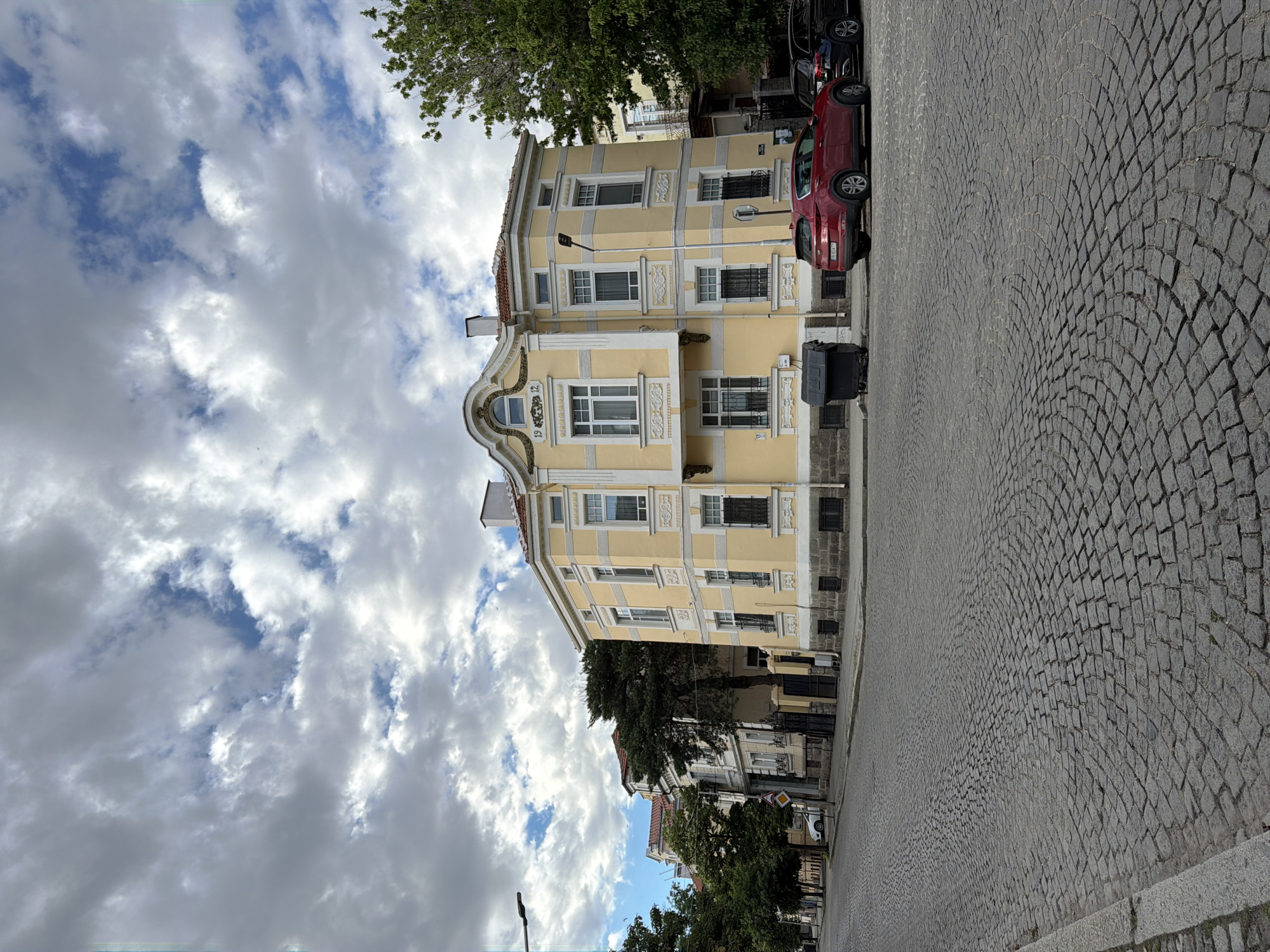 The image depicts a street scene in Plovdiv, Bulgaria, specifically in the area of the Old Town or a similar historic district, evidenced by the cobblestone street and the architectural style of the buildings. The foreground is dominated by a curved cobblestone road, with a granite curb on the left. On the right side of the street, a light yellow, two-story building with ornate white trim and a red-tiled roof is prominently featured. This building has multiple windows with black shutters and decorative elements above them. The number "19 12" is visible above a central window on the upper floor.
Adjacent to this main building, further down the street, are other buildings with similar historical architectural features, some with red-tiled roofs and lighter facades. Trees line the street on the left, and a tall, dark evergreen tree stands between the yellow building and the adjacent structures.
Several cars are parked on the street: a red SUV facing away from the camera is parked in front of the yellow building, and a dark-colored sedan is parked behind it. A large black trash bin is located on the sidewalk in front of the yellow building.
The sky above is filled with dramatic, textured clouds, suggesting a partly cloudy or overcast day. A streetlamp is visible on the left side of the frame, extending over the sidewalk. The scene appears to be a quiet street with historical character.