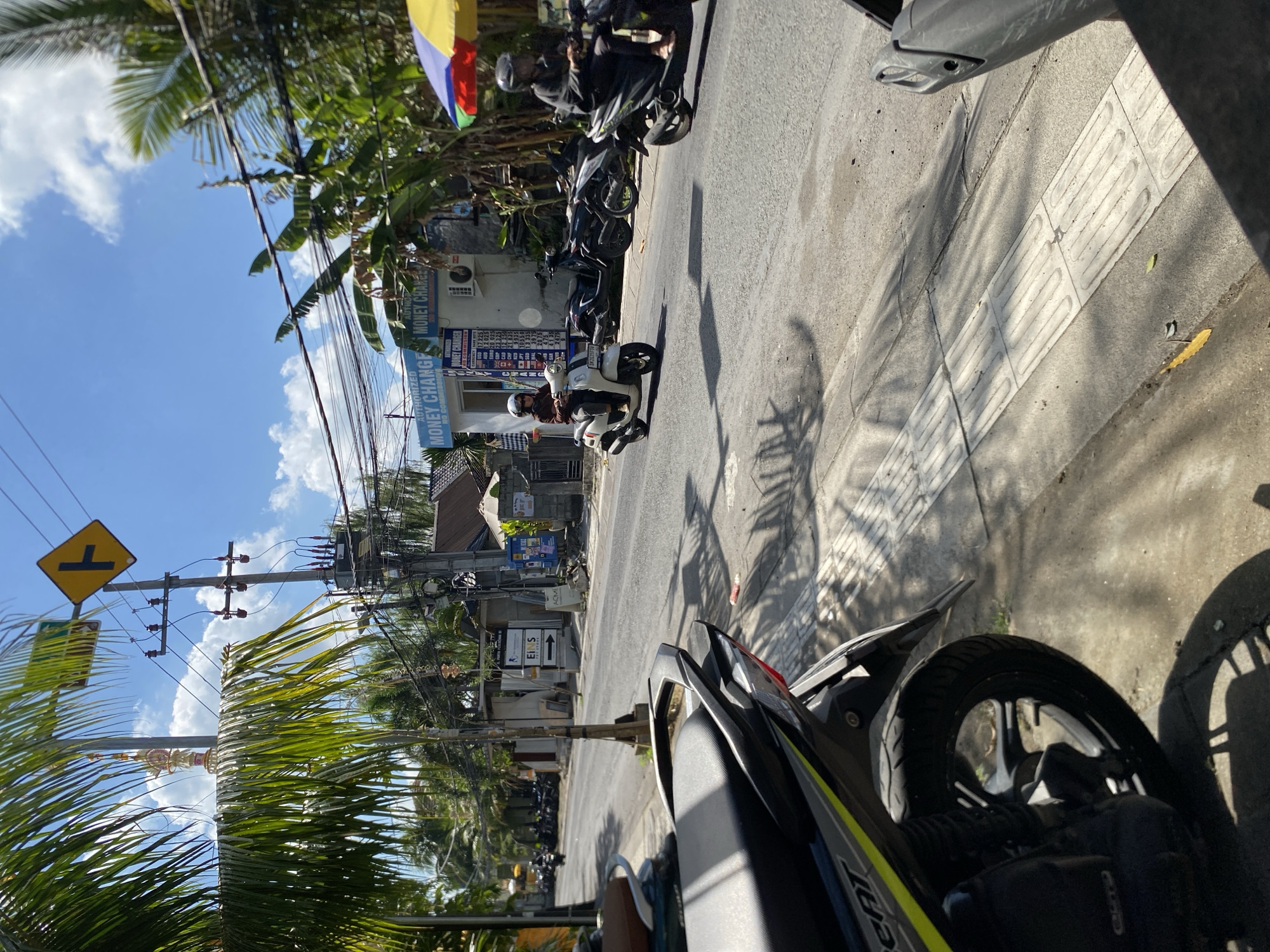 This is a medium-angle, eye-level, outdoor, daytime photograph taken on a street in Mengwi, Indonesia. The scene depicts a road with traffic and buildings lining one side. In the foreground, the rear section of a parked dark-colored scooter is visible on the right. On the left, another parked scooter is partially visible. The road itself is asphalt, with a textured concrete sidewalk running along the right side. The sidewalk has tactile paving for visually impaired pedestrians.
In the mid-ground, a white scooter carrying a person wearing a helmet and sunglasses is traveling away from the camera on the right side of the road. Another scooter, ridden by a person wearing a helmet, is traveling in the same direction further ahead on the right. On the left side of the road, several scooters are parked in front of a building with signage that reads "AUTHORIZED MONEY CHANGER". Above the money changer, more signs and electrical wiring are visible, suspended from utility poles.
The background shows more buildings, palm trees, and other tropical vegetation. The sky is bright blue with scattered white clouds. A yellow diamond-shaped traffic sign with a black T-shaped symbol is visible on a utility pole in the upper left portion of the image. Another religious-looking decorative structure is mounted high on a pole near the traffic sign. The overall impression is of a sunny day on a typical street in a tropical urban or semi-urban area.