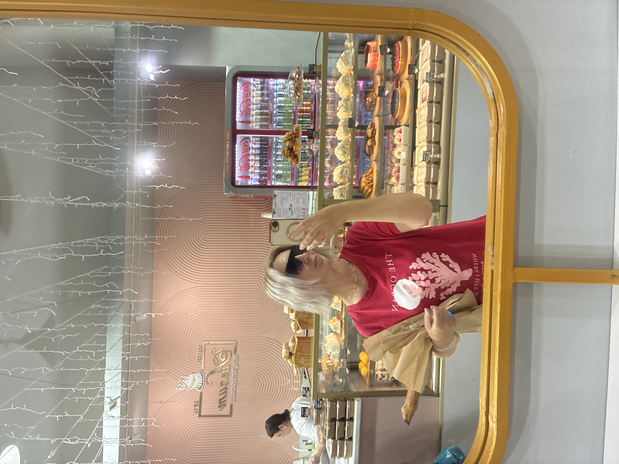 A woman with blonde hair and sunglasses, wearing a red t-shirt with a coral design, is taking a selfie in a mirror. She is holding a baguette wrapped in brown paper. Behind her, a bakery display case is filled with various pastries, cakes, and drinks. A Coca-Cola refrigerator is visible in the background, stocked with bottles. Another woman, wearing a white t-shirt, is seen in the background, working behind the counter. The bakery has a pastel pink, textured wall with a gold logo. The overall scene is well-lit, with string lights hanging from the ceiling. The bakery is located in Sochi, Russia.