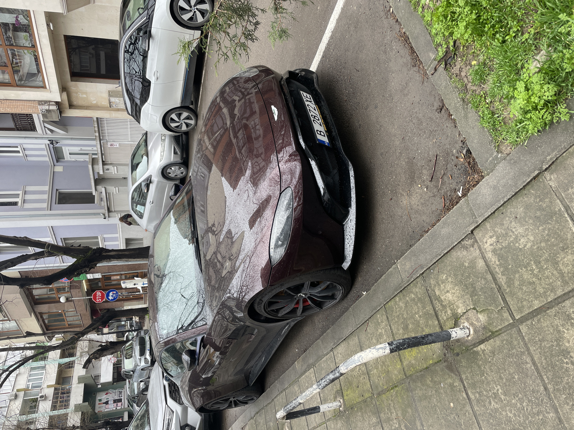 The scene is a street in Varna, Bulgaria, with a dark red Aston Martin parked along the curb. The car's license plate reads "B 2872 XE". It is parked parallel to the curb on a grey concrete sidewalk. To the left of the Aston Martin, several other parked cars are visible, including white cars. Buildings are visible in the background with grey, white, and purple facades. Trees with bare branches line the street. Street signs are visible, including a stop sign. The weather appears overcast, as the car's surface is wet, possibly from rain. The sidewalk adjacent to the curb is cracked and worn, and a black and white painted metal barrier runs along the edge. Some green vegetation is growing in the ground alongside the sidewalk. The perspective is a slightly elevated angle.