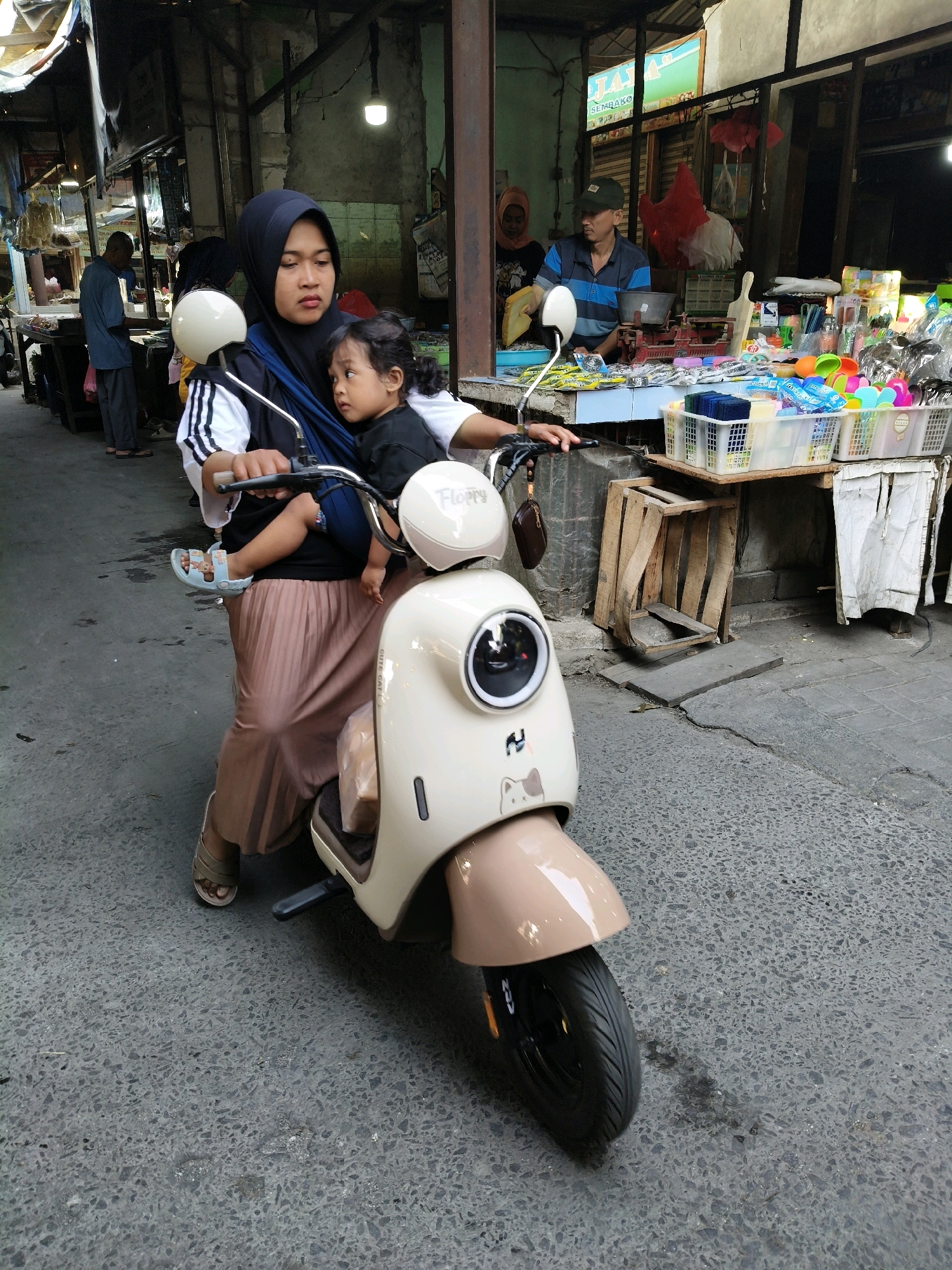In Setiamekar, Indonesia, a woman wearing a black hijab and a beige pleated skirt is riding a cream-colored scooter. She is carrying a young child in a black baby carrier, who is facing away from the viewer. The woman is looking forward with a neutral expression. The scooter has a white headlight with a circular reflective surface and a small cream-colored fin. The background depicts a market scene with stalls selling various goods, including what appear to be household items and food. In the background, a man wearing a blue and black striped shirt and a dark baseball cap is visible behind a stall. The ground is paved with asphalt and shows some signs of wear and tear. The overall environment suggests a busy outdoor market area.