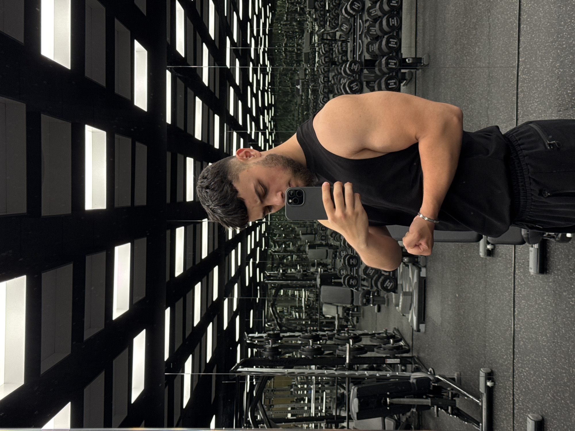 A young man with short, dark, curly hair and a beard is taking a mirror selfie in a well-lit gym. He is wearing a black sleeveless shirt and black athletic pants. The gym has a dark ceiling with a grid of recessed white lights and mirrors lining the walls, reflecting rows of weightlifting equipment, including dumbbells and benches. The flooring is dark and speckled. He is holding a smartphone in his right hand, angled to capture his reflection. His left arm is flexed, showcasing his bicep. The overall impression is of a modern fitness facility. There is no indication of the city of Varna, Bulgaria, in the image itself.