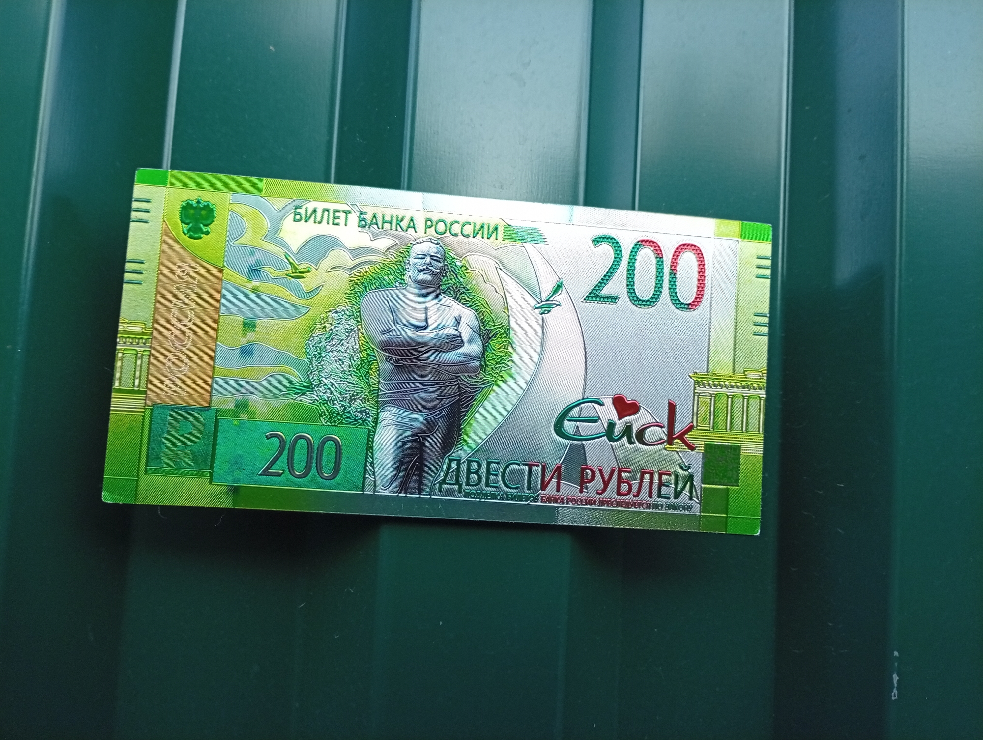 A 200 Russian ruble banknote is partially visible, lying on a green corrugated metal surface. The banknote features a depiction of a statue of a man with his arms crossed, and architectural elements. The denomination "200" is clearly visible in large green numerals. The background is a textured, dark green metal surface with vertical ridges. No people are present in the image. The location of Pavlovsky Posad, Russia is not depicted or suggested by any elements in the image.