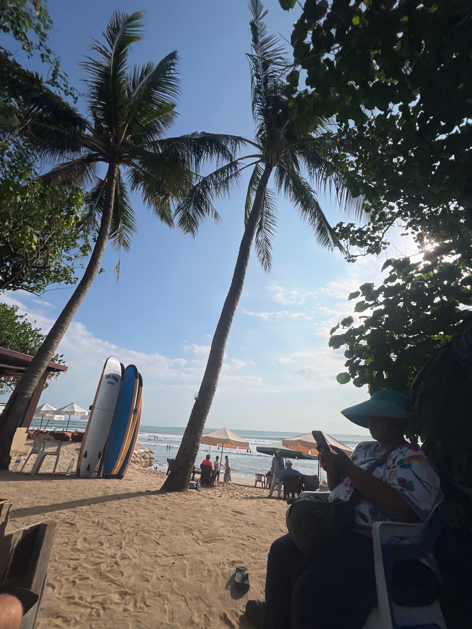 This is a medium shot of a sunny beach scene in Kuta, Indonesia, with two prominent palm trees framing the shot. In the foreground, a person wearing a wide-brimmed hat and a floral shirt is seated in a chair, looking at their phone. Behind them, on the sandy beach, are several surfboards leaning against each other. Further back, people are seen relaxing under beach umbrellas, and some are in the ocean. The sea and a cloudy sky are visible in the background. The lighting suggests it is late afternoon.