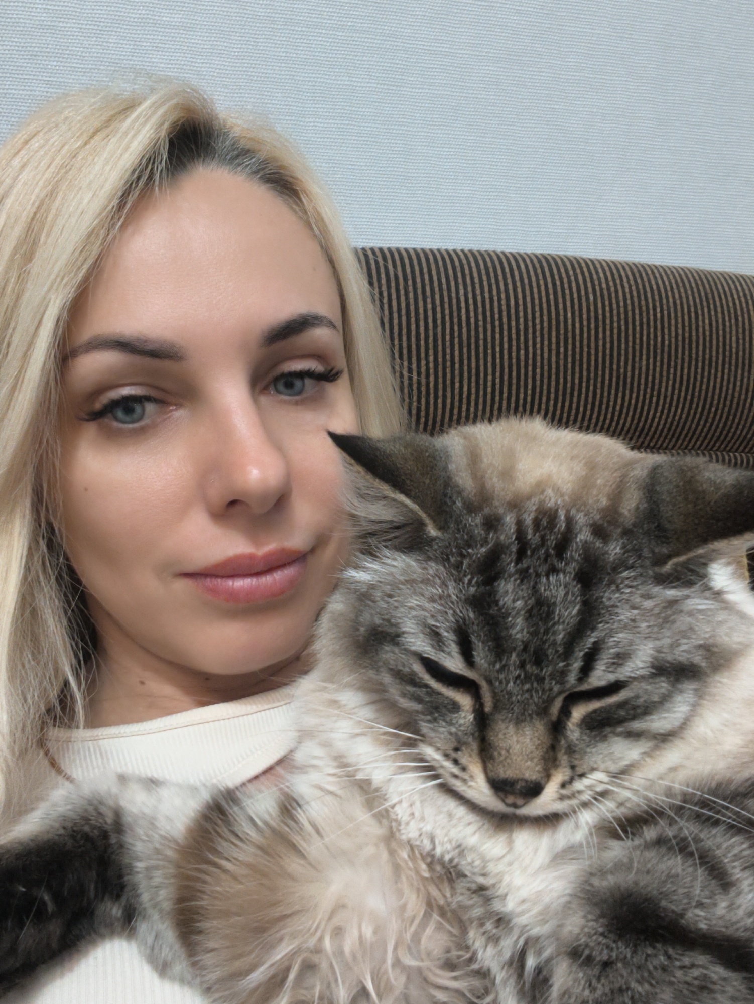 A blonde woman with blue eyes is holding a sleeping cat. The woman is wearing a light-colored shirt. The cat is gray and beige. They are sitting on a brown and black striped couch. The background is a light blue wall. There is no indication of the location in the city of Sevastopol, Ukraine.