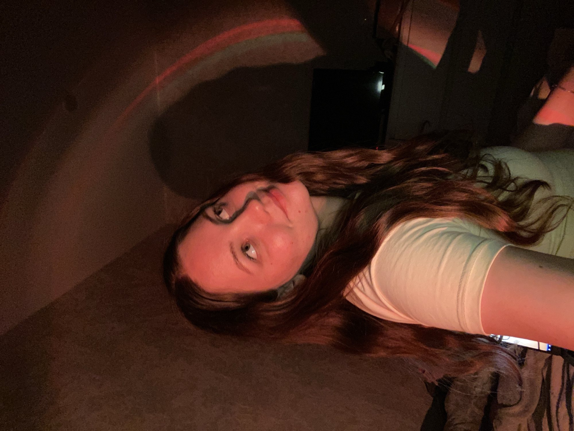 The image shows a young woman with long, wavy brown hair, looking upwards and to her right. She is wearing a light-colored t-shirt. The scene is dimly lit, with a reddish hue cast over her face and hair, possibly from a colored light source. In the background, there is a dark wall and a faint outline of what appears to be a television or monitor. A shadowy, arched light projection is visible on the wall above and behind her. The overall environment suggests an indoor setting, possibly a bedroom, with soft furnishings visible in the lower left corner. There is no indication of the specific location being in Izhevsk, Russia.