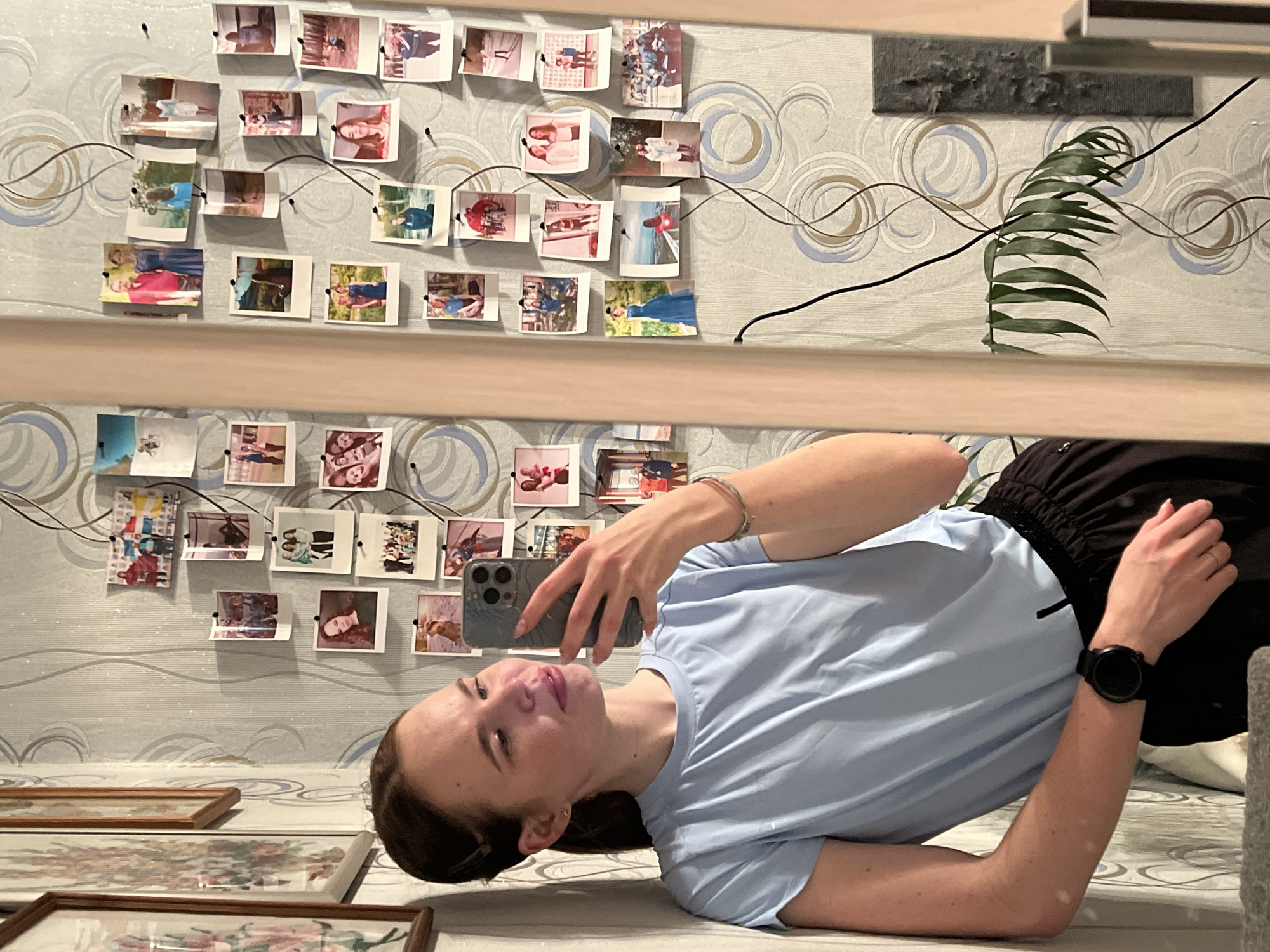 The image shows a young woman lying on a bed in a room. She is holding a smartphone in her left hand, seemingly taking a selfie. She is wearing a light blue t-shirt, black shorts, and a black smartwatch on her right wrist.
The room has wallpaper with a pattern of circles and curved lines. A collection of photographs is pinned to the wall above the bed, arranged in two rows. A framed picture hangs on the lower left of the bed frame. There is a green plant behind the frame. The location is Chaikovsky, Russia.