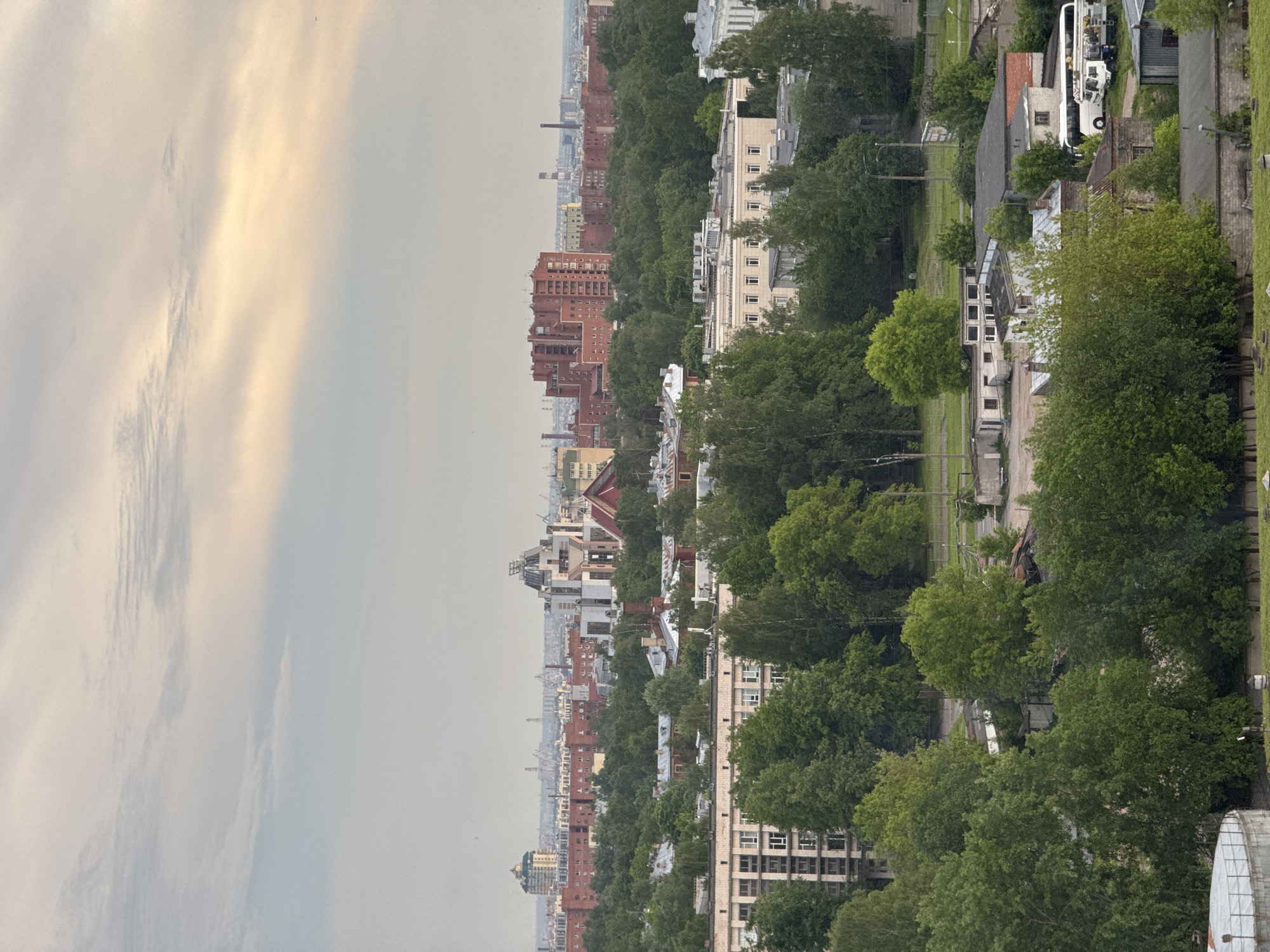 The image displays an elevated panoramic view of a city or town under an overcast sky. The original image is rotated 90 degrees clockwise, presenting a landscape scene in portrait orientation.
In the corrected landscape view:
The upper portion of the frame shows a soft, overcast sky with varying shades of grey and muted yellow, suggesting diffused sunlight breaking through or reflecting off clouds.
The midground and foreground are dominated by a dense canopy of lush green trees, likely from parks or tree-lined streets, interspersed with various urban structures. A prominent feature is a series of red-brick buildings, some tall and appearing older, possibly industrial or multi-story residential, stretching across the middle distance. Alongside these are several lighter-colored, cream or white buildings, possibly apartment blocks or commercial buildings, with numerous windows.
Further to the right, nestled among trees and smaller buildings, a large white passenger bus is visible. Pathways or roads can be glimpsed winding between the foliage and structures. In the far distance, the city extends into a hazy horizon, with more indistinct buildings and what appears to be a tall industrial chimney fading into the atmospheric haze.
The scene depicts a calm daytime atmosphere. The weather is overcast, and the distant haze suggests a potentially humid or atmospheric condition. There are no people visible, and no specific activities are taking place beyond the general urban setting. No discernible text is present in the image. The architecture hints at a possible European or post-Soviet urban environment, but no definite location cues are present.