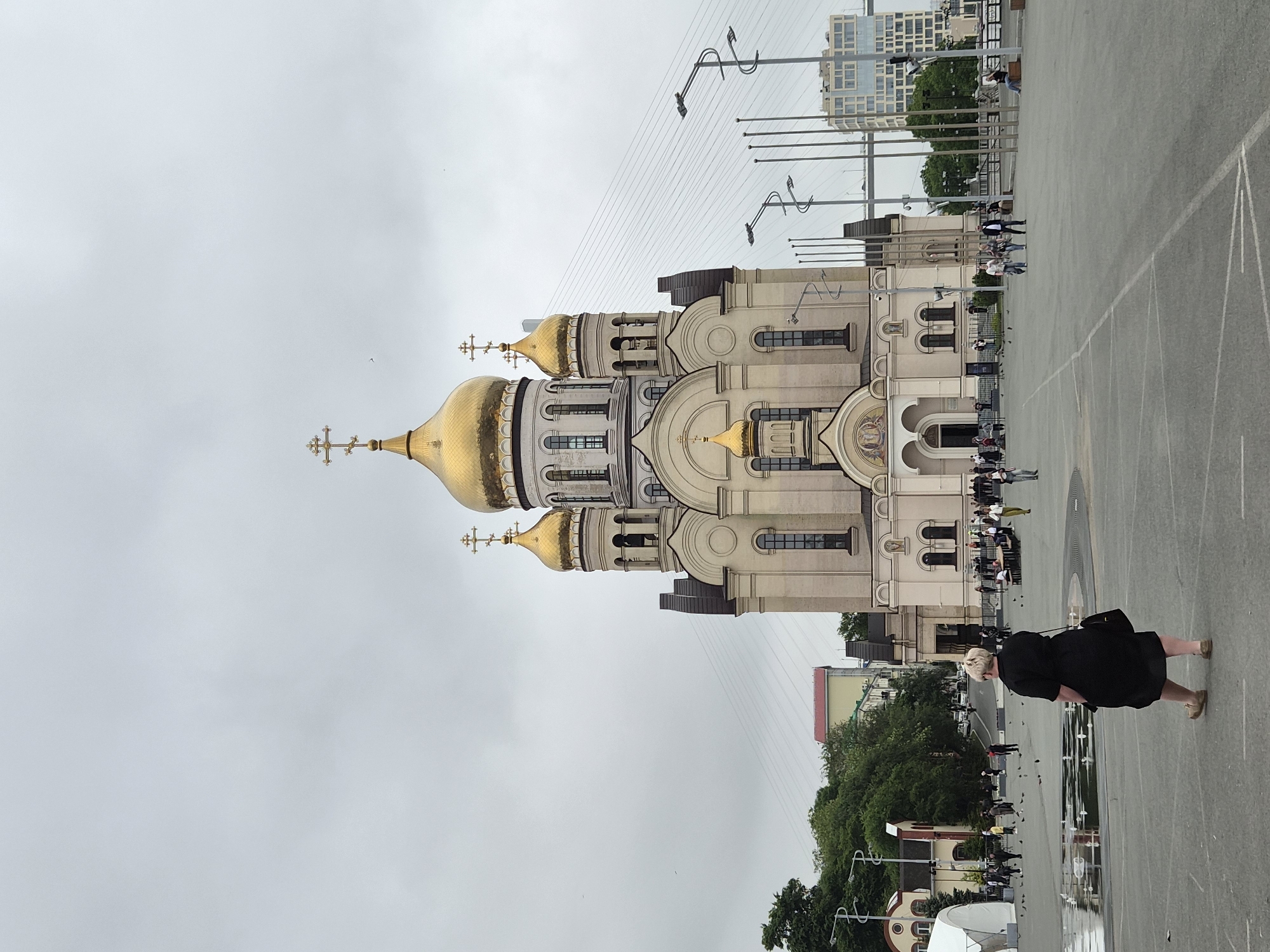 A wide-angle, eye-level, outdoor shot captures the Church of the Assumption of the Virgin Mary, a prominent landmark in Vladivostok, Russia, under an overcast sky. The church, with its distinctive gold domes and intricate architecture, dominates the center of the frame. In the foreground, a woman with blonde hair wears a black dress and carries a dark handbag. She is seen from behind, walking away from the camera towards the church. Several other people are scattered across the plaza, some walking, others standing or sitting. A small group is gathered near tables and chairs in front of the church. Pigeons are present on the plaza, with some walking near a shallow pool of water on the left side of the image. In the background, trees, buildings, and the suspension cables of the Golden Horn Bay Bridge are visible. The overall atmosphere is serene and somewhat muted due to the cloudy weather.