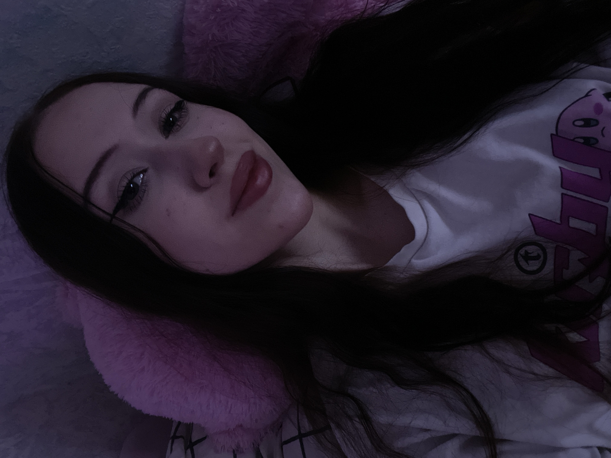 This is a close-up, high-angle, selfie-style portrait of a young woman with long, dark hair. She is lying down, with a pink, fluffy pillow behind her head. The woman is wearing a white t-shirt with a graphic print, possibly a cartoon character. She has on makeup, including eyeliner and lipstick. The lighting is dim, with a purple hue, suggesting it was taken indoors at night or in a dimly lit environment. There is no discernible information about the location in Votkinsk, Russia, or any specific context beyond what is visually present in the image.
