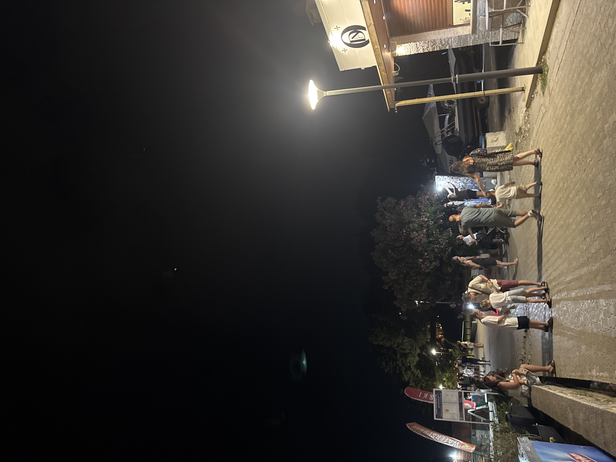 This image depicts a nighttime scene with a group of people gathered on a paved walkway. The time of day is clearly night, indicated by the dark sky and the artificial lighting. The walkway appears to be near a body of water, possibly a harbor, as boats are visible in the background. The setting suggests a public area, perhaps a waterfront promenade or a street adjacent to a marina.
Several individuals are present, some standing and conversing, while others are seated or leaning. They are dressed in casual summer attire, suggesting a warm climate and possibly a social gathering. The mood appears relaxed and convivial. A street lamp illuminates the area, casting a glow on the people and the surroundings. In the background, there is a building with signage, partially obscured by darkness and foliage.
Visible text includes a sign with "SKI" and "WATERSPORTS" which, along with the boats, strongly suggests a recreational area, likely related to water activities. The presence of multiple boats in the background and a sign advertising water sports points to a coastal or lakeside location. The overall impression is one of a lively evening, perhaps a market or a gathering place for tourists and locals alike. The ground is wet, possibly indicating recent rain or a cleaning effort, which reflects the artificial lights.