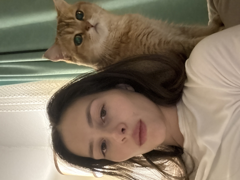 An adult female and a domestic cat are depicted in an indoor, close-up shot. The female, positioned in the foreground, has dark hair, fair skin, and is wearing a white garment. Her eyes are directed towards the camera. To her right and slightly behind her, a golden-orange colored cat with prominent emerald green eyes is visible, also looking towards the camera. The background features a light-colored, patterned curtain or wall covering on the left, and a solid teal-colored curtain or fabric on the right. A warm light source is partially visible on the far left. The subjects appear to be situated within a domestic environment. There are no visual cues within the image to confirm a specific location such as Yekaterinburg, Russia.