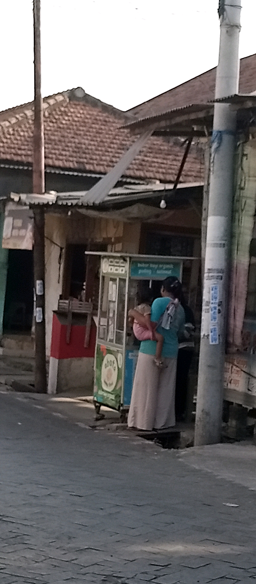A medium-shot, slightly blurred, outdoor scene in Wadungasri, Indonesia, shows a woman standing in front of a food cart, holding a toddler. The woman, wearing a teal shirt and a long beige skirt, has the toddler on her front. The food cart is primarily green and white, with signage that is difficult to read. Behind the cart, a red and white structure is visible. A utility pole stands to the right of the scene, adorned with various posters. The background features a building with a tiled roof and corrugated metal awnings. The ground is paved with bricks, and the sky is overcast.
