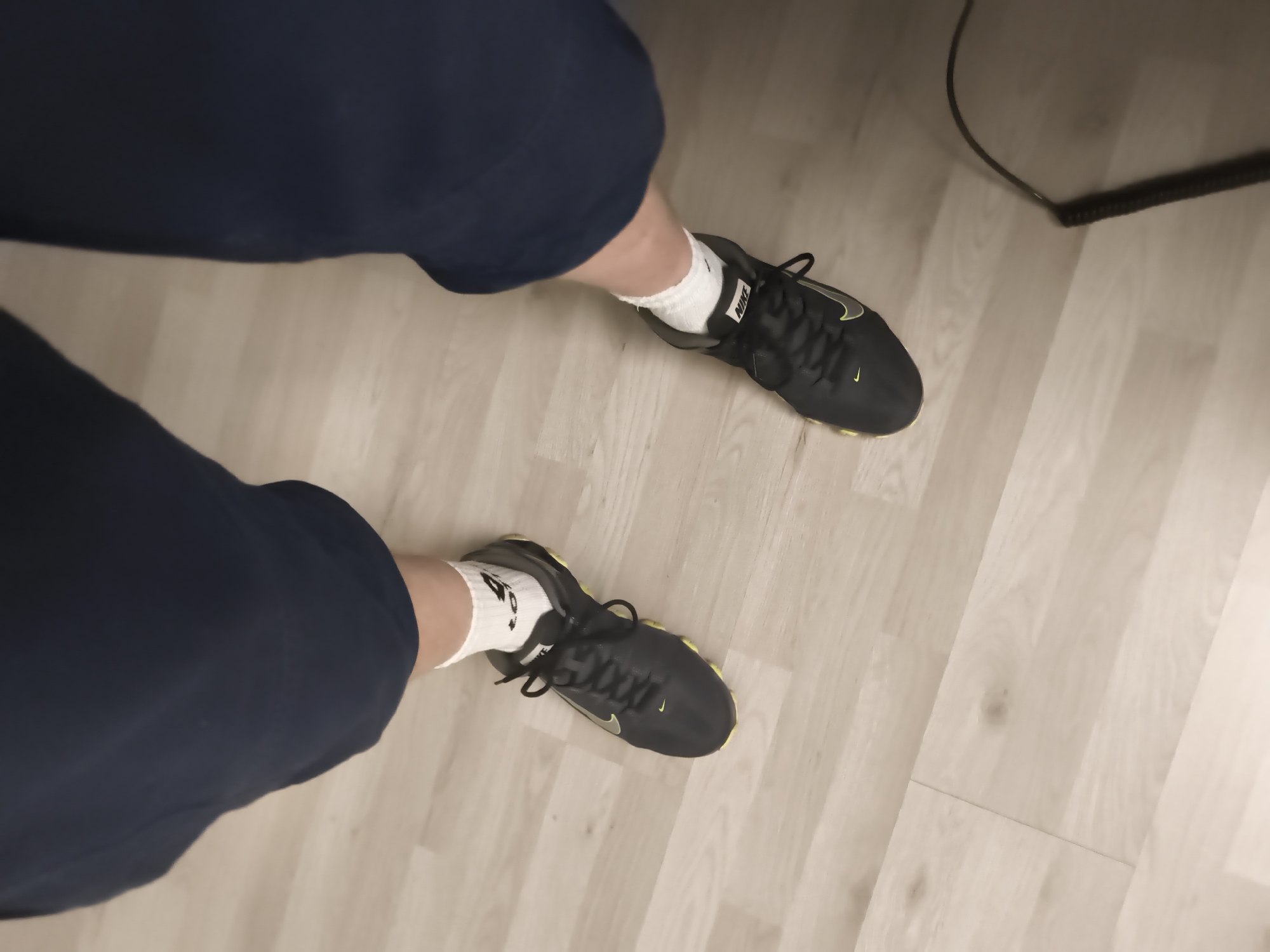 The image shows a low-angle view of a person's legs and feet, wearing dark blue pants and white athletic socks with black Nike shoes. The shoes are primarily black with a neon green swoosh logo and accents. The person is standing on a light-colored wooden floor. In the upper right portion of the image, a black cord, possibly from an electronic device, is visible on the floor. The setting appears to be indoors, given the flooring and the presence of the cord. There is no indication of the time of day or weather conditions, and no discernible emotions are conveyed. The text "NIKE" is visible on the side of the shoes, and a small Nike swoosh is also present on the toe area of both shoes. The location context is Troisdorf, Germany.