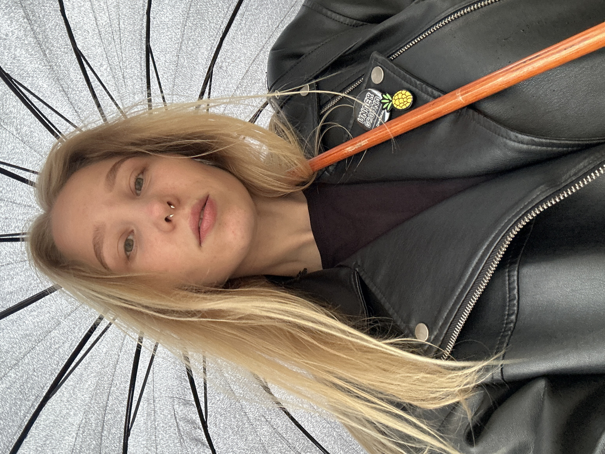 A woman with light blonde hair, a nose ring, and fair skin looks up at the camera from under an umbrella. She wears a black leather jacket over a black top. A wooden umbrella handle is visible across the jacket. A round pin with a pineapple is on the lapel, next to a rectangular pin with the words "UNDER GROUND" in a stylized font. The location is not identifiable from the visual information provided.