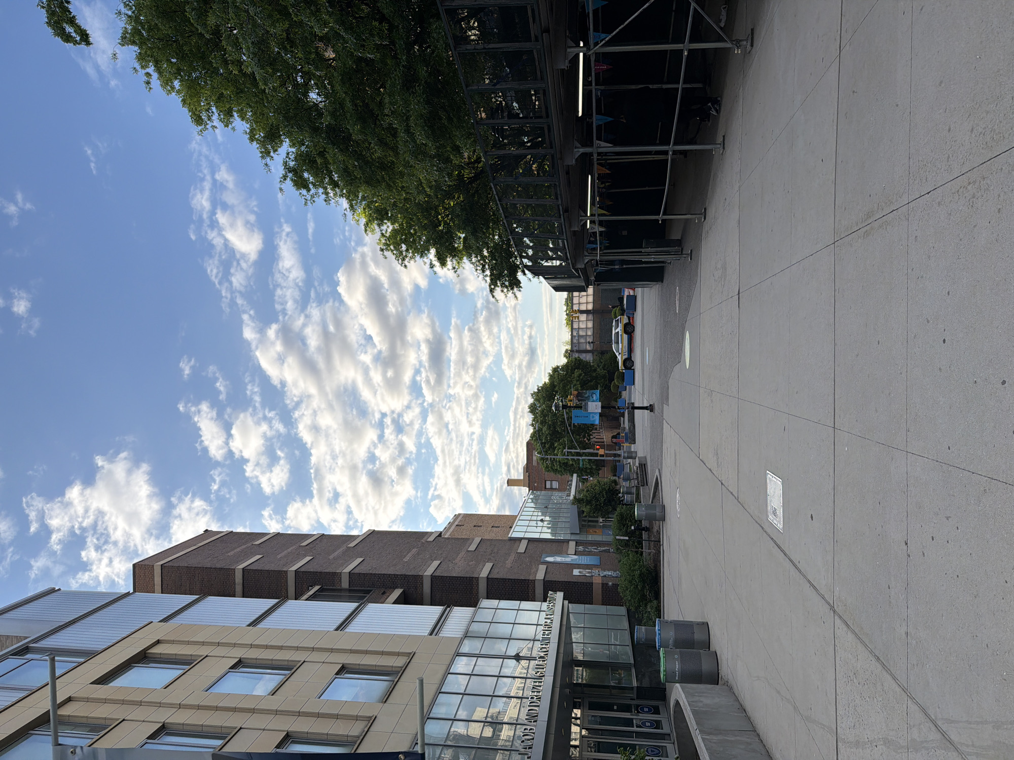 The image shows a street scene in New York City, United States of America. The Jacob and Drexel Galuch Shulmann museum is visible in the foreground. The sky is blue with some fluffy white clouds. There are some trees visible, and also some scaffolding along the street. The road is a smooth paved road.
