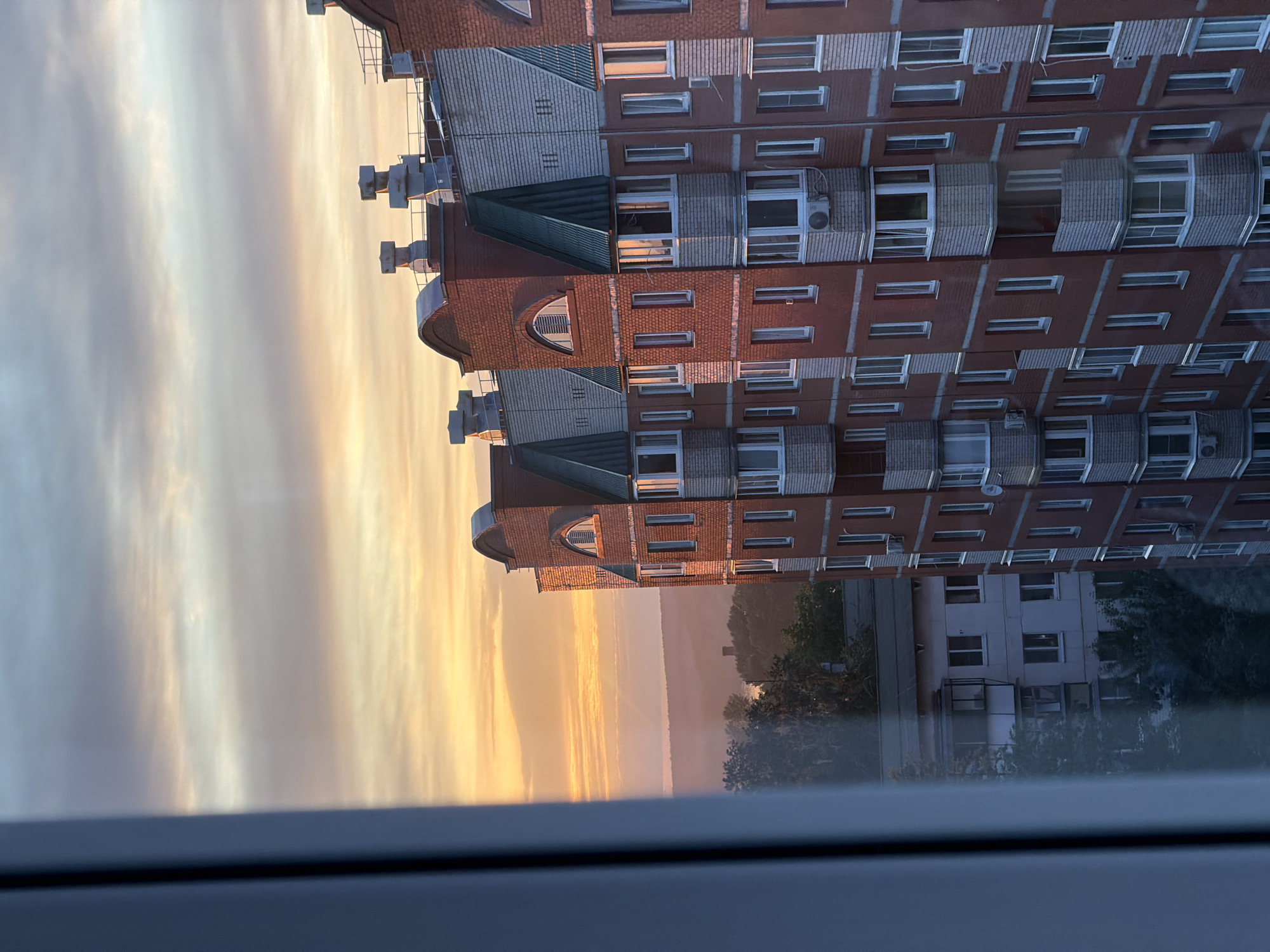 The image shows a view from a window in what appears to be Chaikovsky, Russia, based on the architectural style of the buildings. The foreground is the window frame, partially obscuring the view. Looking out from the window, a multi-story brick building is prominent on the right side of the frame. The building is red brick with white window frames and some windows have air conditioning units. The building has a distinct architectural style. To the left of the red brick building is the edge of the lake Kama. The sky is filled with clouds, and the sun is setting (or rising). The colors are soft, with hues of yellow, orange, and gray. Another building is visible in the distance.
