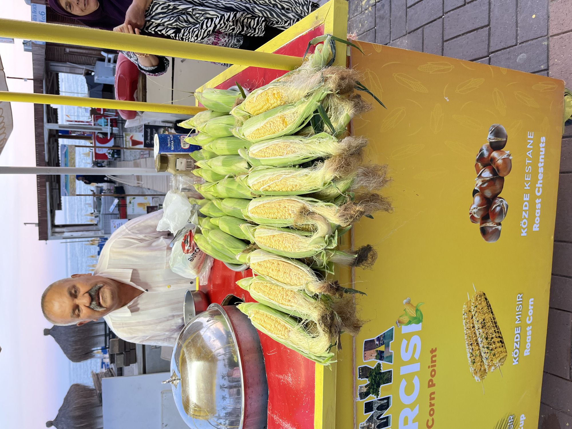 This image depicts a vibrant street food stand, likely located on a promenade or beachside in Alanya, Turkey. The central subject is a smiling man with a prominent mustache, dressed in a striped shirt, who appears to be the vendor. He stands behind a yellow and red cart. Another person, a woman wearing a patterned top and a dark hijab, is partially visible in the upper left corner, looking towards the camera.
The stand is piled high with fresh corn on the cob, many with their green husks partially peeled back, revealing bright yellow kernels and silken tassels. A large, gleaming silver domed pot, likely used for cooking the corn or chestnuts, is prominent on the red section of the cart. Next to it are a smaller silver bowl and a white plastic bag, with the word "BUTCH" partially visible. A small blue container labeled "İYOTLU SOFRA TUZU" (Iodized Table Salt) from the brand "BİLSA" is also seen.
The side of the cart features clear signage. Text readable includes "ANTALYA RCISI Corn Point," indicating the type of business and a regional connection to Antalya. Key menu items are advertised as "KÖZDE MISIR Roast Corn" and "KÖZDE KESTANE Roast Chestnuts," accompanied by illustrative images of corn and chestnuts.
In the background, a body of water, likely the sea, is visible with beach umbrellas and distant buildings under a soft, possibly late afternoon or early evening sky. The ground is paved with grey interlocking tiles. The scene evokes a casual and lively atmosphere typical of a coastal resort town.