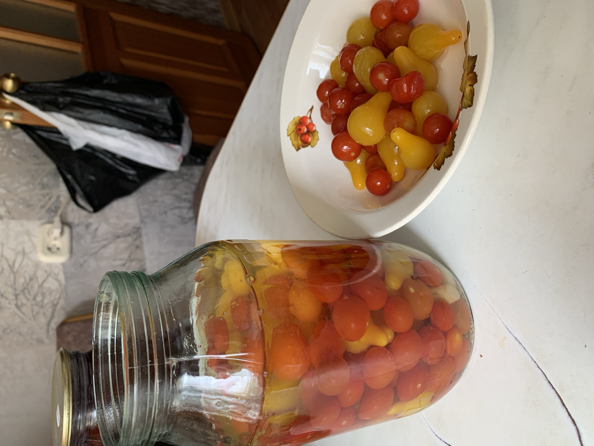 A high-angle shot shows a table with a jar of pickled tomatoes and a bowl of fresh tomatoes.
The jar, made of clear glass, is lying on its side and contains a mix of red and yellow tomatoes submerged in a brine. The lid is visible and appears to be metal.
Beside the jar is a white bowl filled with a similar mix of red cherry tomatoes and yellow pear-shaped tomatoes. The bowl features a fruit design on its side.
The table is white and there are other objects in the background, including a black trash bag. The location is identified as Chaikovsky, Russia. The overall impression is of a kitchen or pantry scene where food is being prepared or preserved.