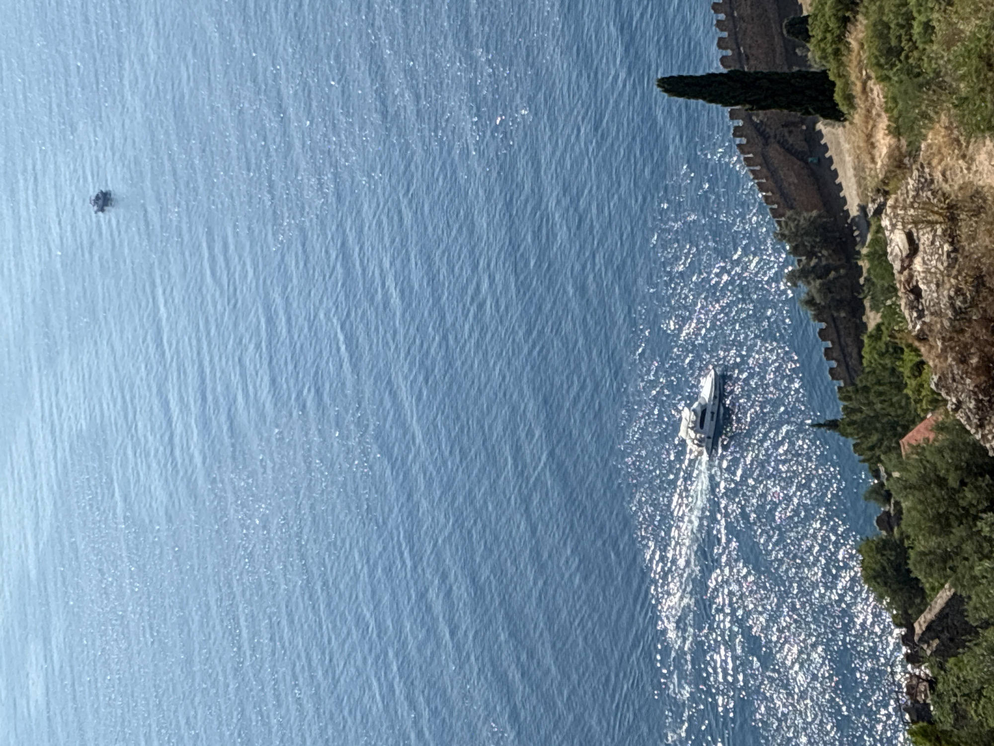 This is an aerial view of the Mediterranean Sea from the Alanya Castle in Alanya, Turkey. The image shows a vast expanse of blue water with gentle waves, reflecting the bright sunlight. A white motor yacht is visible in the lower left quadrant of the image, moving away from the viewer and leaving a white wake behind it. Further out in the sea, a smaller, dark-colored boat is visible in the upper right quadrant, appearing as a small silhouette against the water. The foreground of the image features the ancient stone walls of Alanya Castle, with its crenellated battlements casting shadows. Green cypress trees and other lush vegetation are also present on the hillside leading up to the castle walls. The scene depicts a sunny day with clear skies, suggesting a pleasant atmosphere for recreation and tourism.