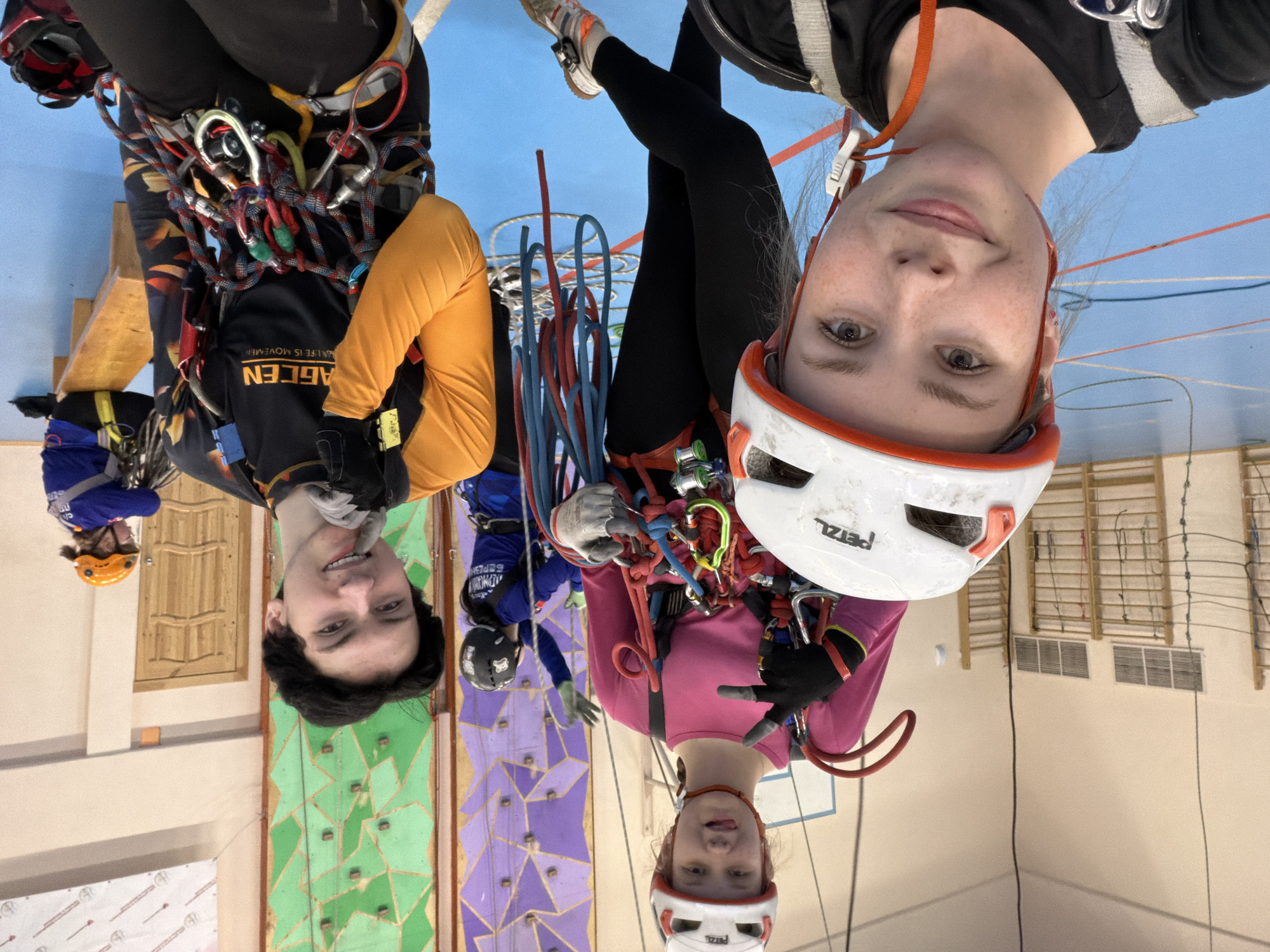 Several individuals are seen in an indoor climbing facility. Three people are the focal point: a male in a black shirt with yellow sleeves and a climbing harness, and two females in helmets, also in climbing harnesses. One female wears a pink top, while the other wears a black t-shirt. The individuals are surrounded by climbing gear and ropes. A climbing wall is visible in the background. The location may be a climbing gym in Berezniki, Russia.