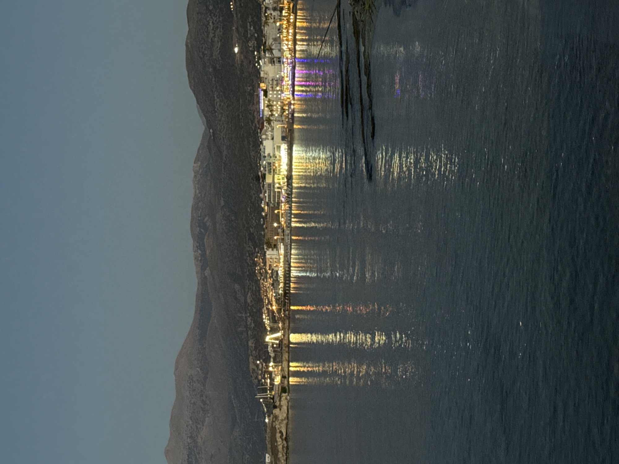 An evening view captures a coastal town with lights reflecting on calm water. The scene is set against a backdrop of dimly lit mountains. The town, situated in the Chersonisos Municipal Unit of Greece, features buildings with illuminated windows and streetlights, creating a vibrant glow. Palm trees are visible along the shoreline, and several boats are docked in the harbor. The water is dark, with shimmering reflections of the town's lights stretching across its surface. Some rocky outcrops are visible near the shore in the foreground. The sky is a deep twilight blue, with no visible celestial bodies.