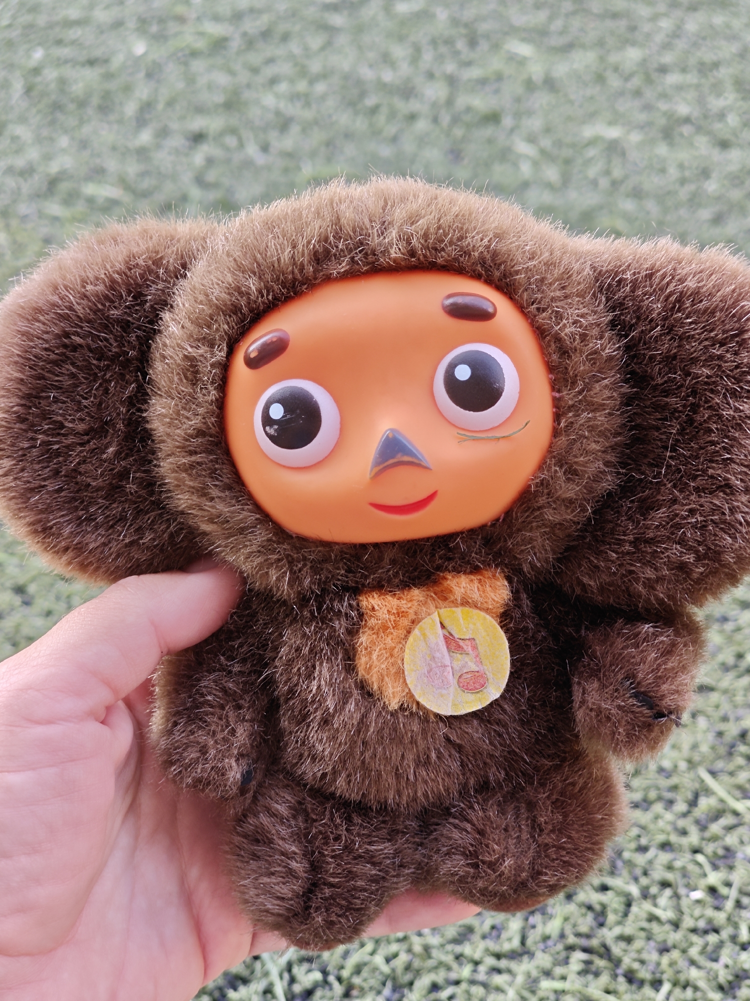 A hand holds a Cheburashka plush toy against a blurred green background. The toy has an orange plastic face with large eyes, a small triangle nose, and a red smile. It is surrounded by brown fur, with large, round ears. A circular tag with musical notes is attached to the toy's chest. The background appears to be artificial turf. The image was not taken in Mumbai, India.