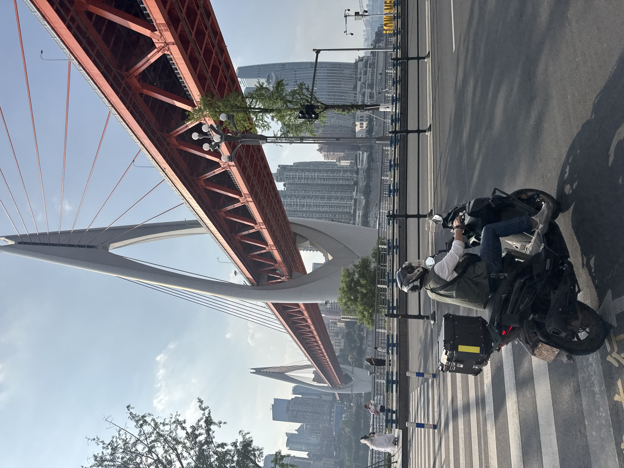 The image shows a person riding a motorcycle on a street in Nan'an District, China. The motorcycle is black and has a storage box attached to the back. The person is wearing a white shirt, blue jeans, and a helmet. The street has a crosswalk painted on it and a bridge in the background. The bridge is red and white and is suspended by cables. There are buildings in the background, as well as a river.